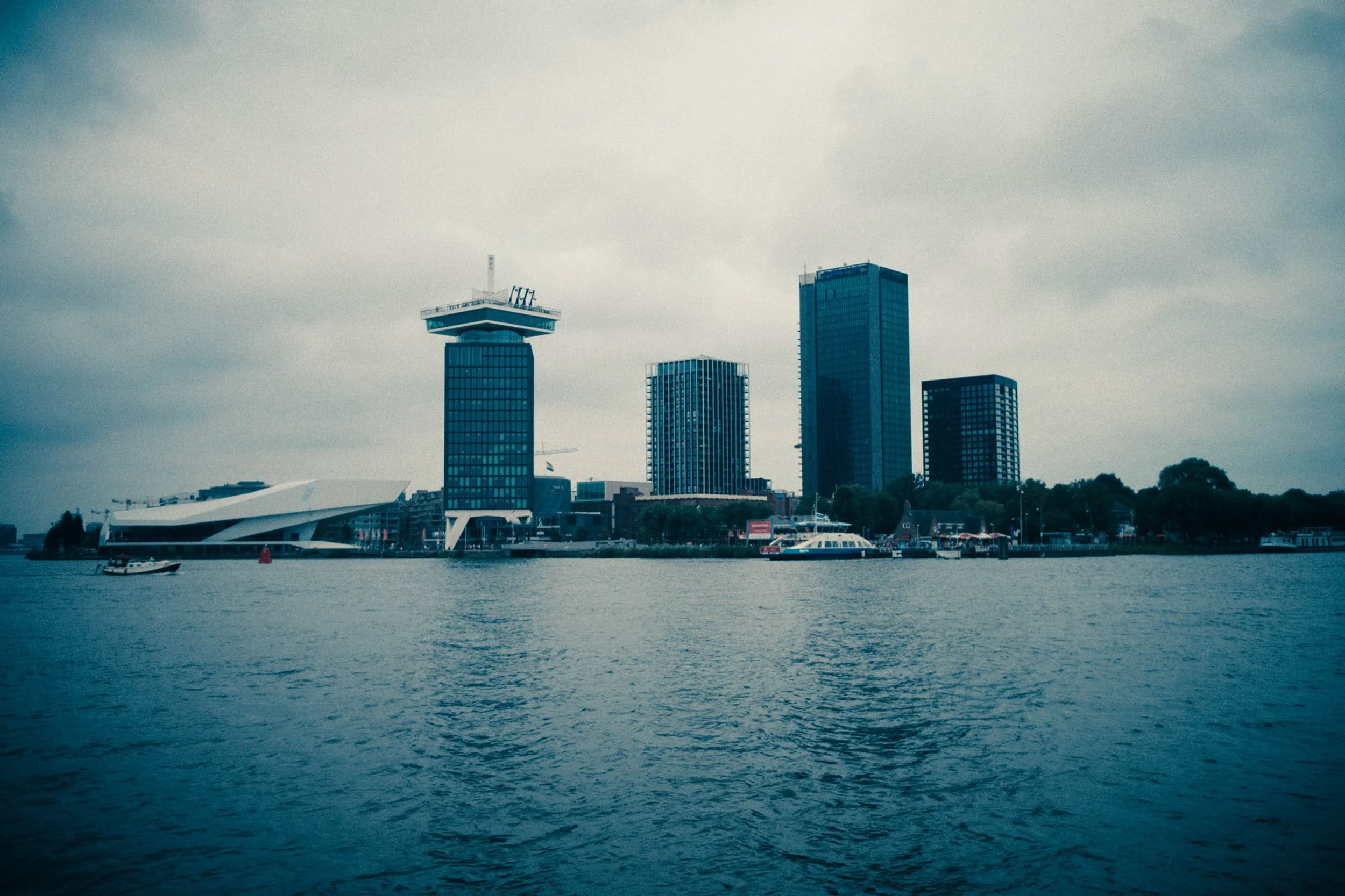 Fotografie van een skyline met hoge gebouwen langs het water, inclusief een modern uitziend gebouw met een opvallend uitkijkplatform, en boten op het water.