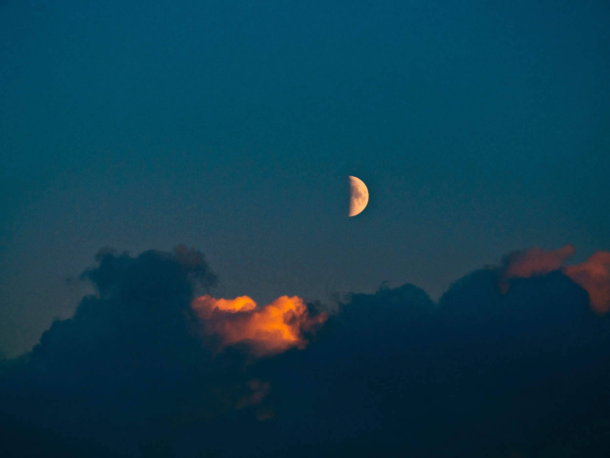 De maan zichtbaar in de lucht tussen donkerblauwe wolken met oranje en roze accenten.