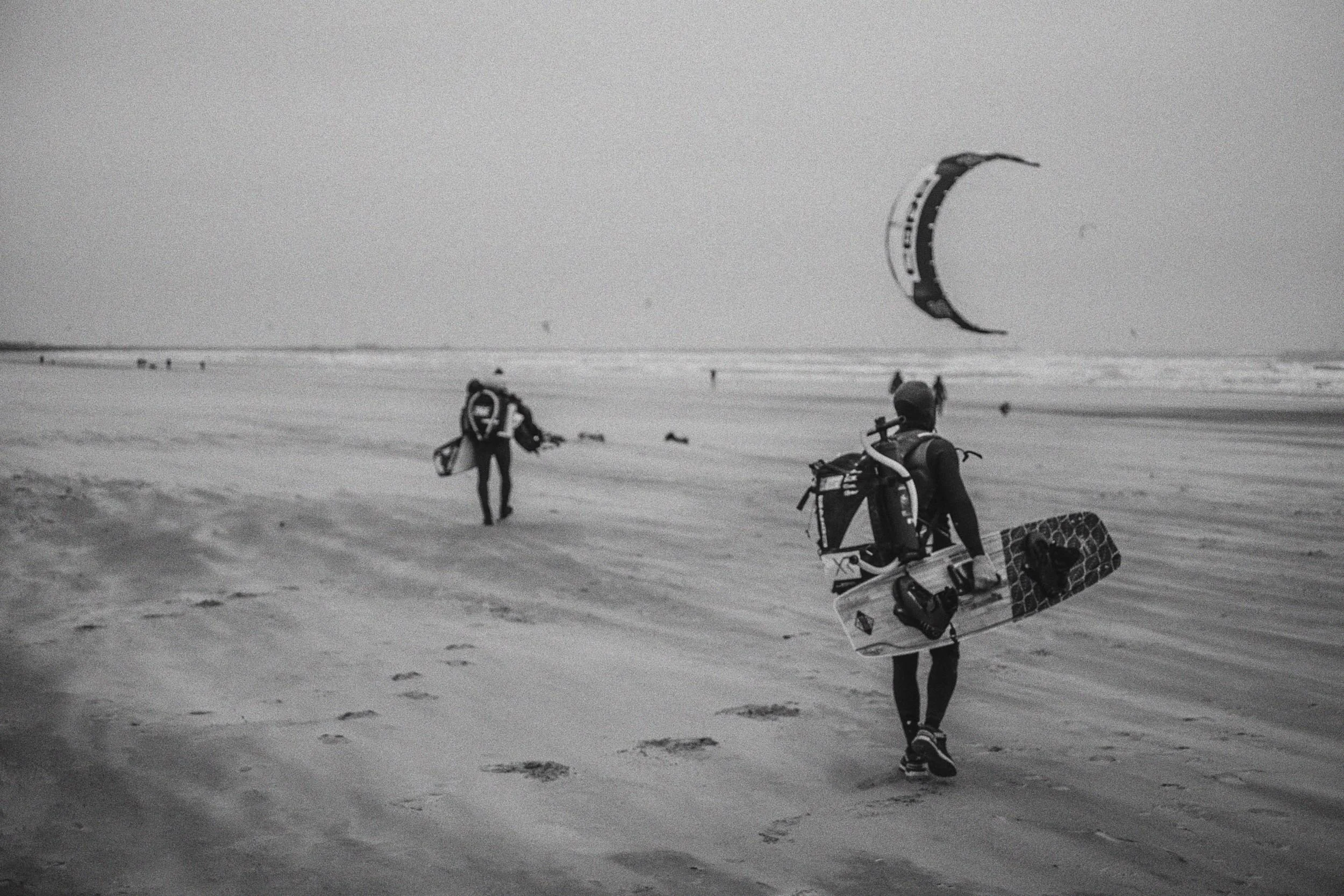 Twee kiteboarders lopen over het zandstrand met hun uitrusting, terwijl een kite in de lucht staat en de zee op de achtergrond te zien is.