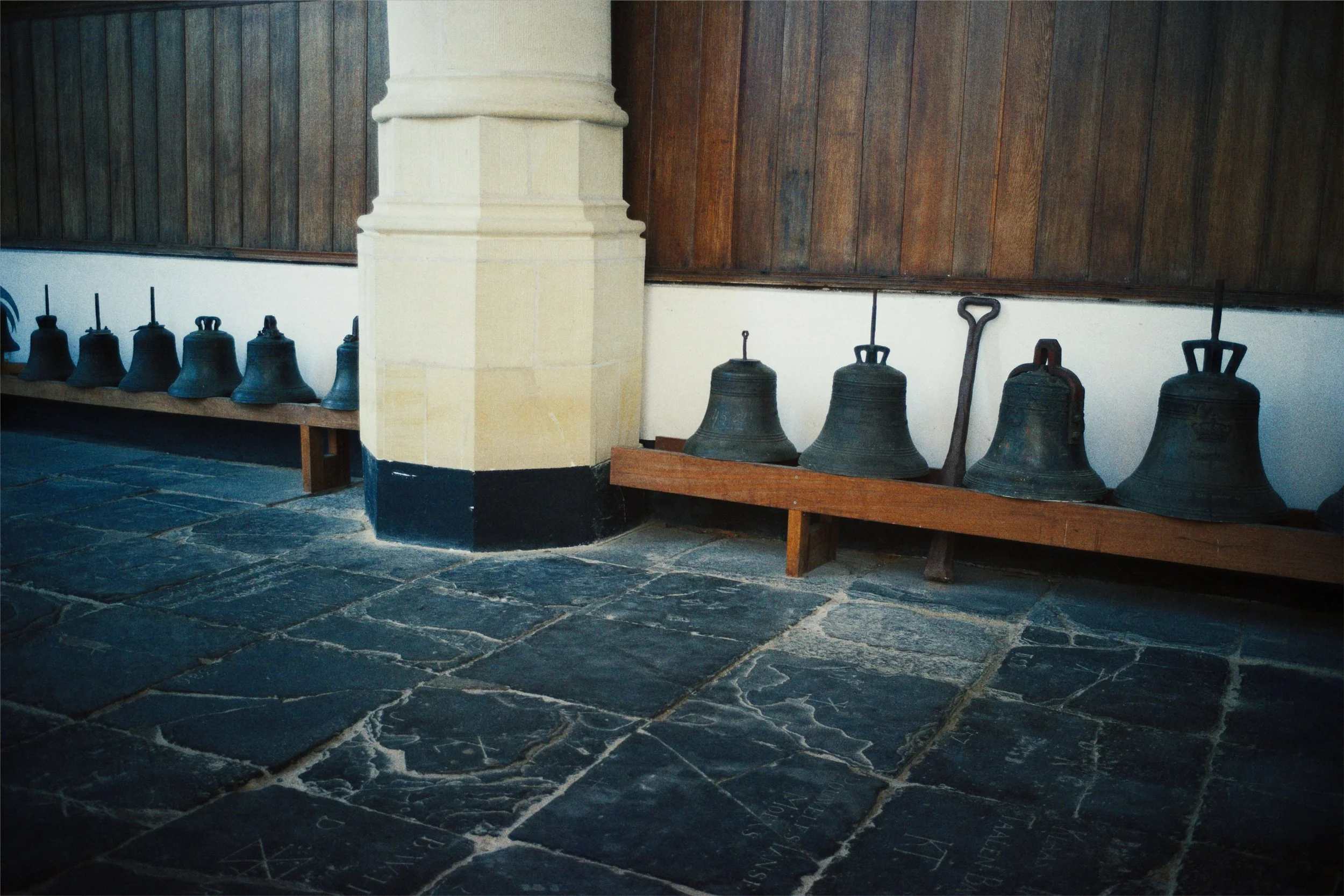 Rijen oude klokken, variërend van klein tot groot, geplaatst op houten rekken langs een muur in een kerk. De vloer bestaat uit grote stenen tegels met inscripties.