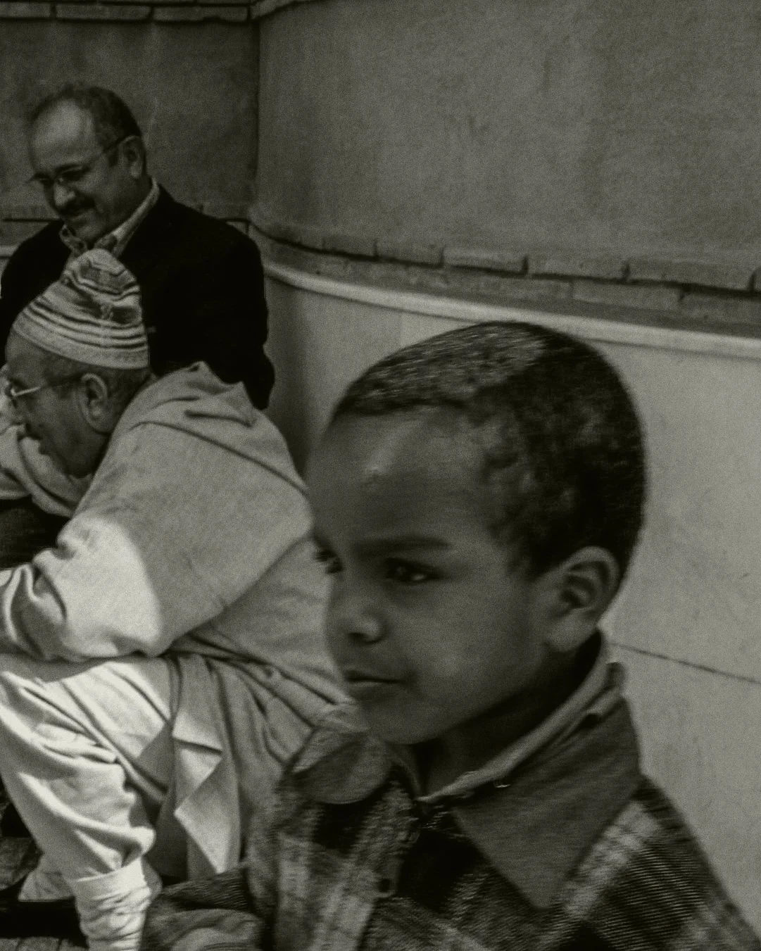 Een zwart-wit foto van een jongen met kort haar en een ruitjes overhemd, zittend en naar rechts kijkend, op de achtergrond staan twee volwassenen die lachen.