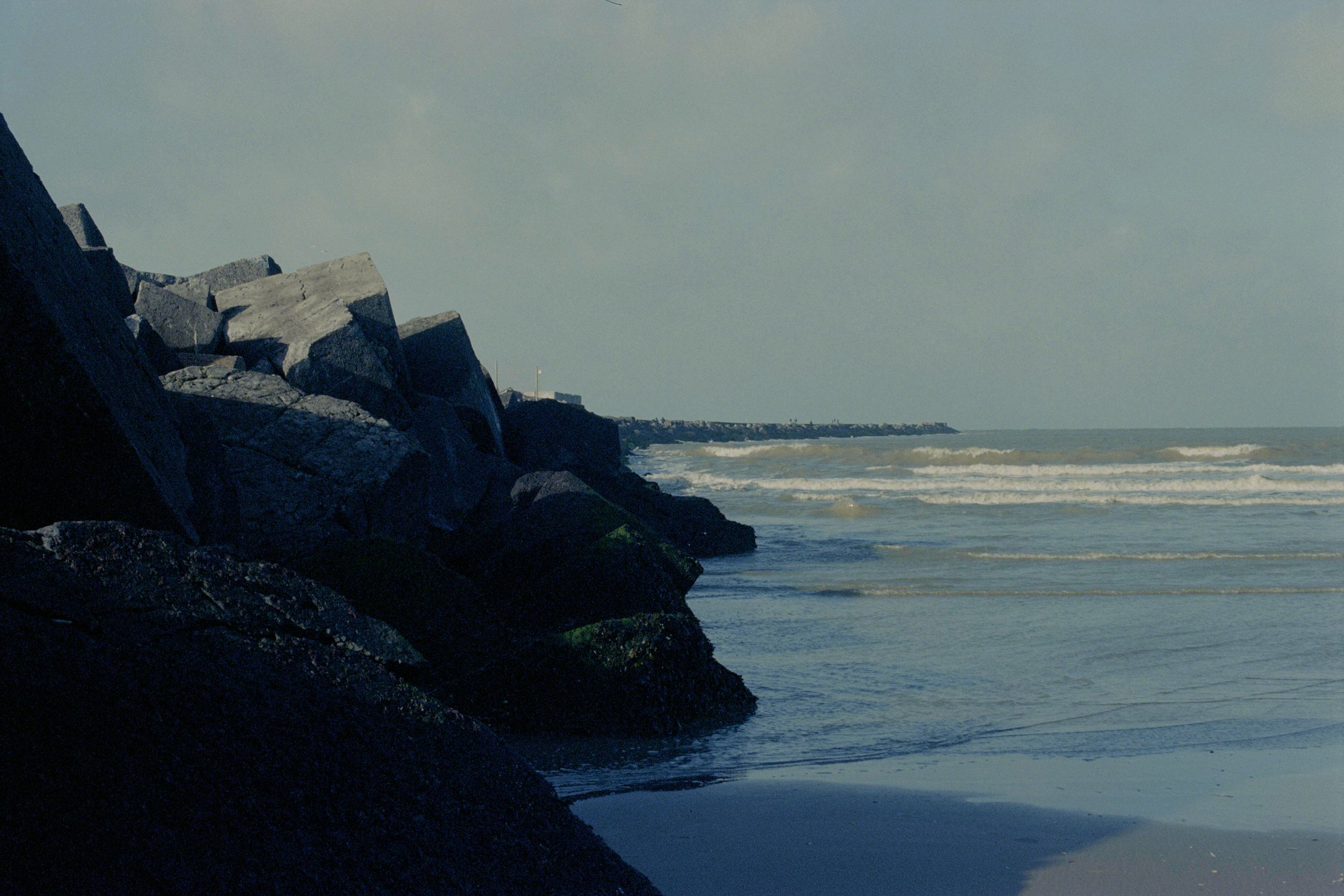 Kust met grote rotsen op de voorgrond en het strand met de oceaan op de achtergrond, onder een bewolkte hemel