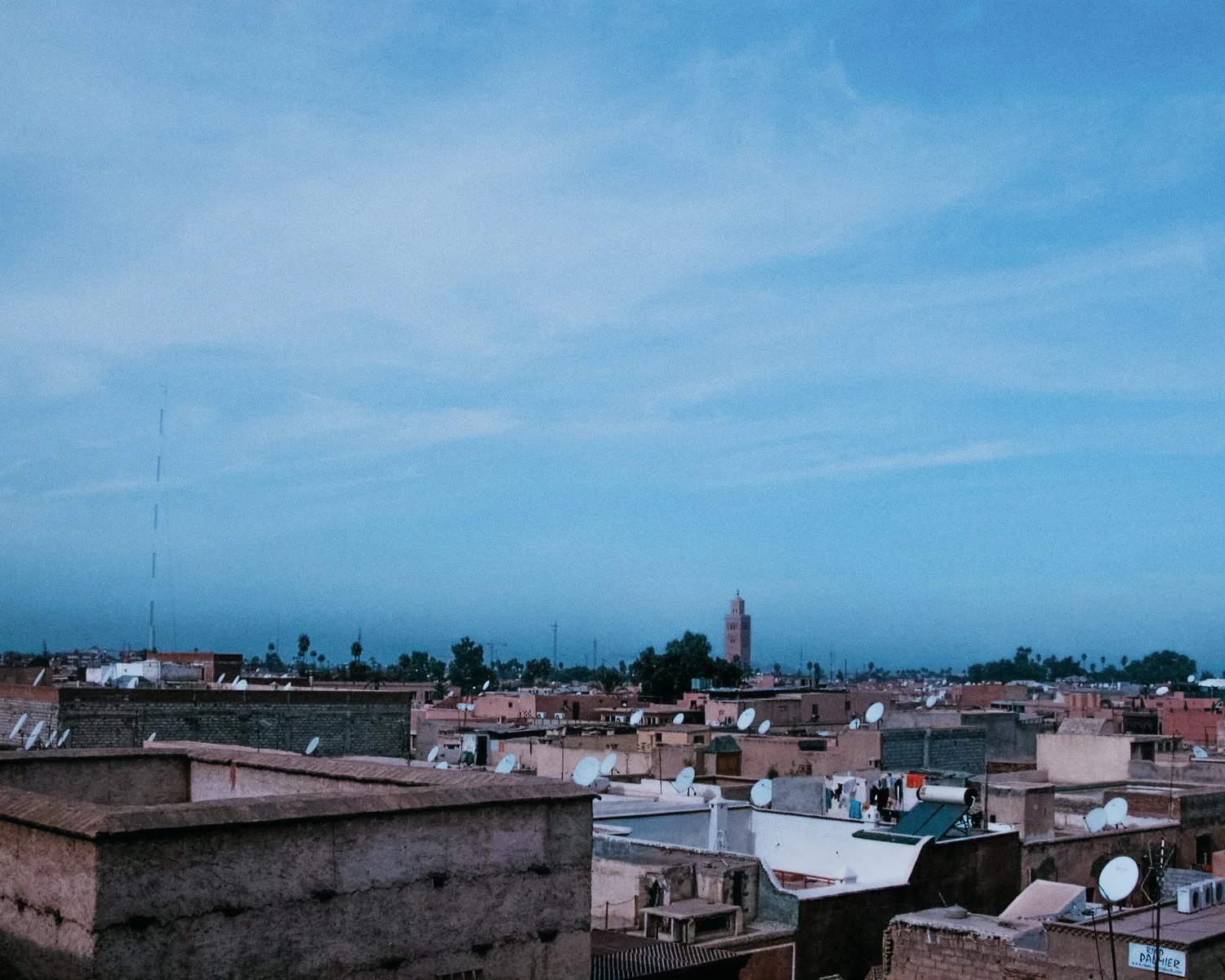 Uitzicht over een stad met platte daken vol satellietschotels, met een blauwe hemel en een lange toren in de verte.