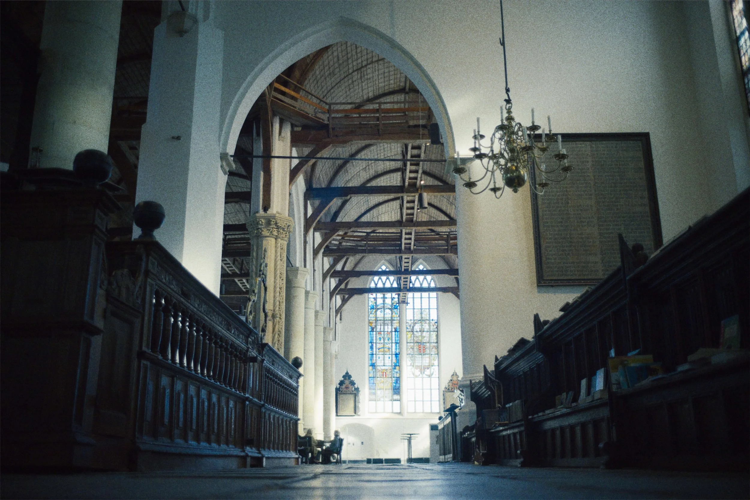Interieur van een kerk met houten bankjes, een grote gekleurde glas-in-loodraam, een kroonluchter hangend aan het plafond, en scheve houten plafondbalken.