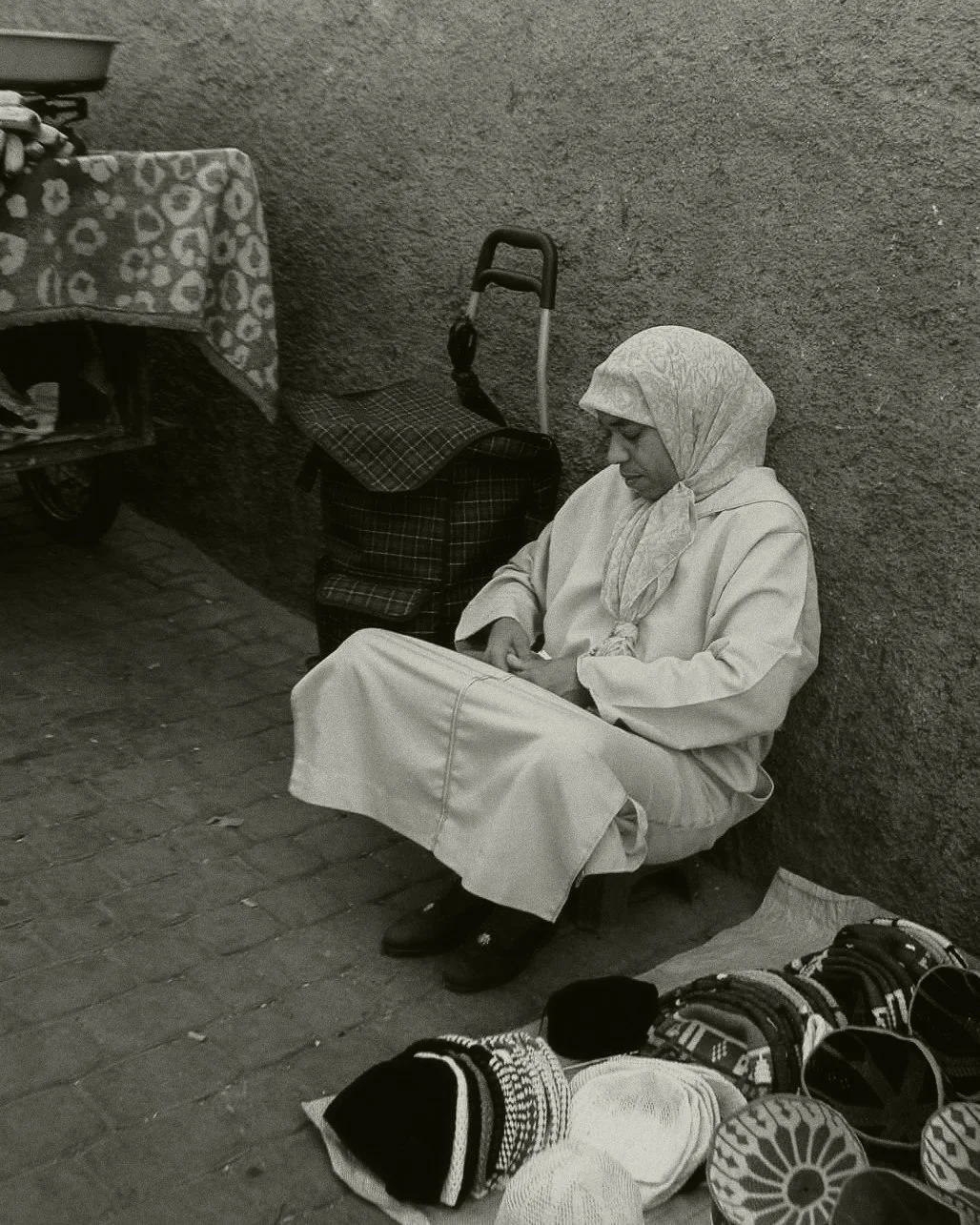Een vrouw met hoofddoek zittend tegen een muur, met diverse hoeden en spullen op de grond voor haar en een wandelwagen naast haar.