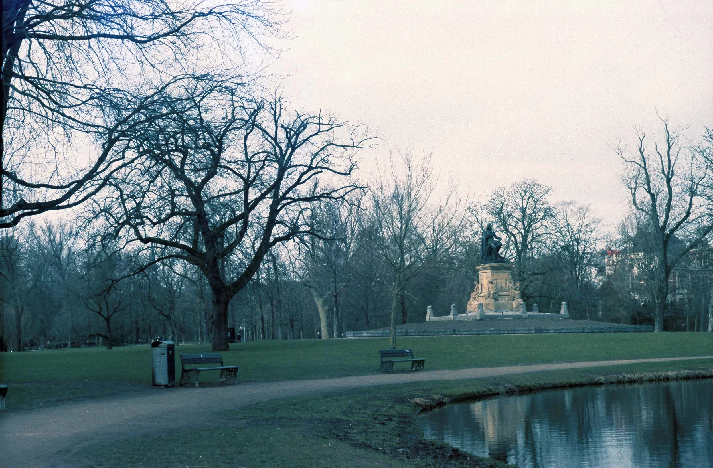 Een park met bomen zonder bladeren, een fontein met een standbeeld, bankjes, een wandelpad en een vijver, op een bewolkte dag.