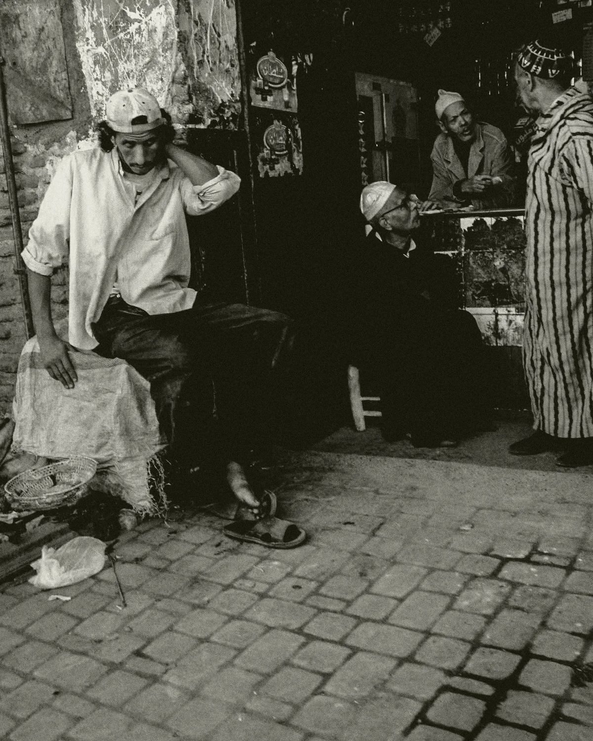Een zwart-wit foto van vier mensen buiten een winkel. Eén man zit op een kruk met een pet achter zijn hoofd en een blouse, terwijl een andere man in een striped outfit en een hoofdband staat. Twee vrouwen zitten in de winkel, een met een hoed en de a