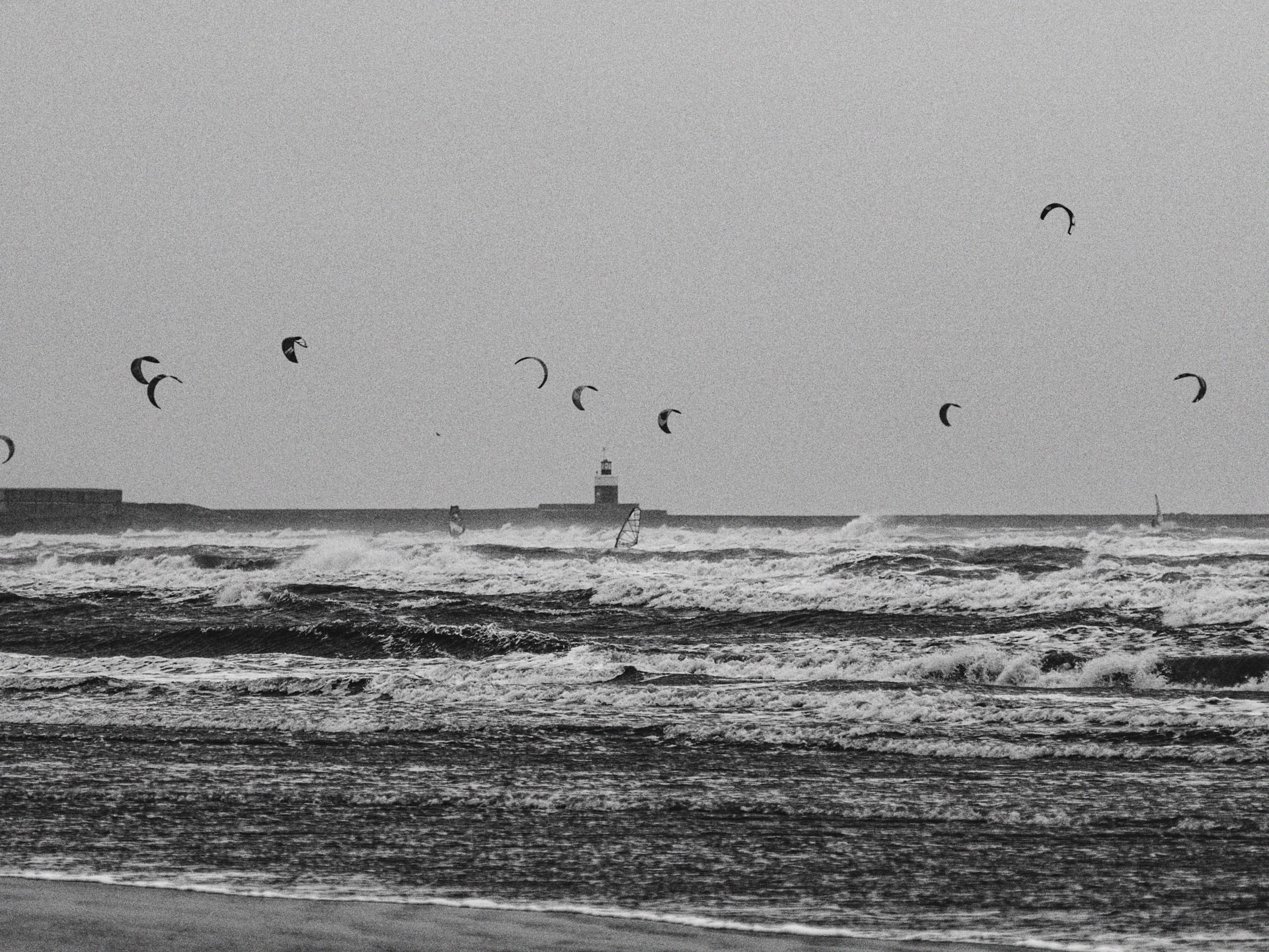 Zwart-wit foto van een drukke zee met surfers en surfers die vliegeren, met een vuurtoren op de achtergrond.
