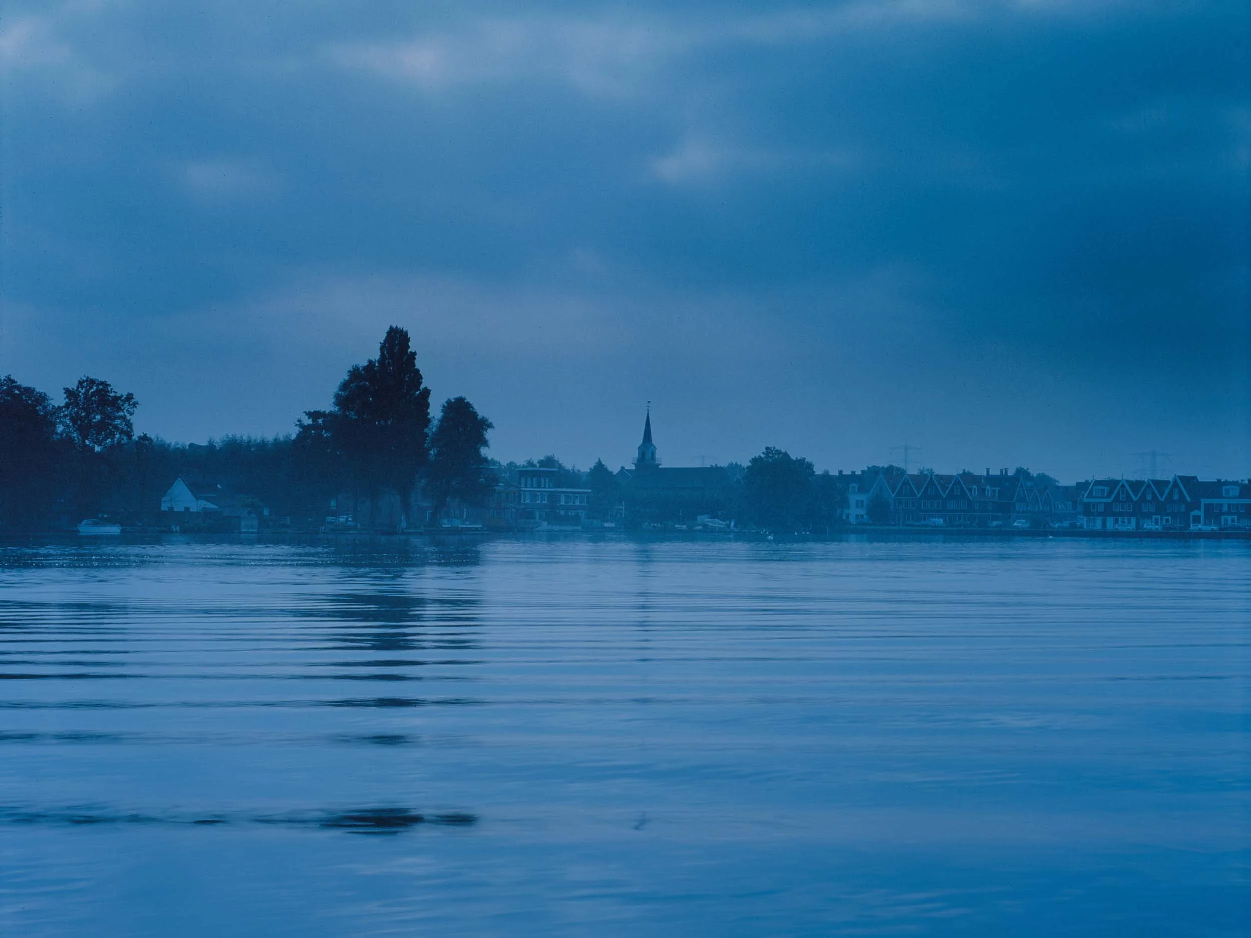 Een rustige rivier met een weerspiegeling van een dorp met huizen en een kerktoren onder een bewolkte hemel.
