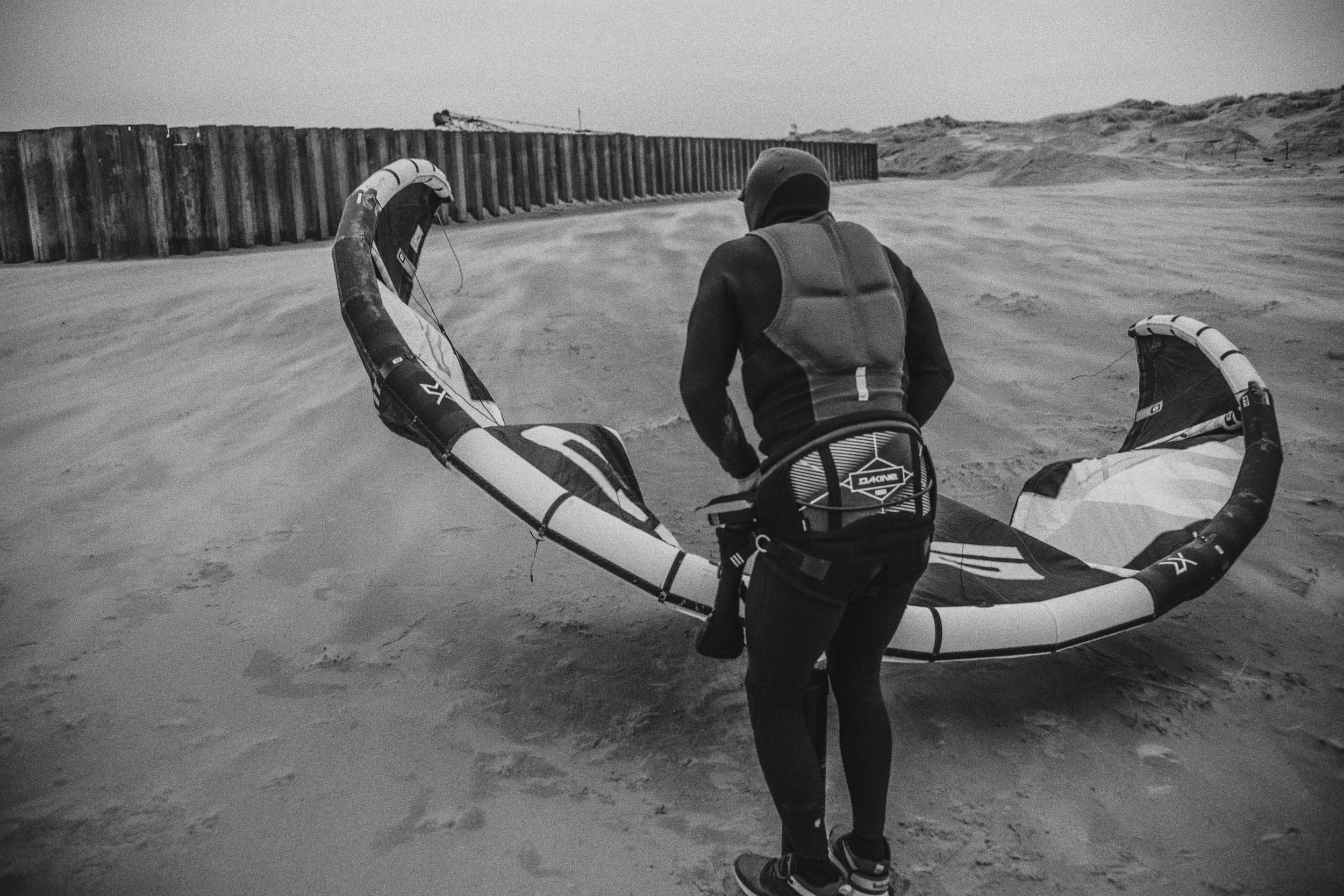Persoon in wetsuit met waterkiter op een zandstrand, met een windgidzend tuig in zn hand, klaar voor watersport.