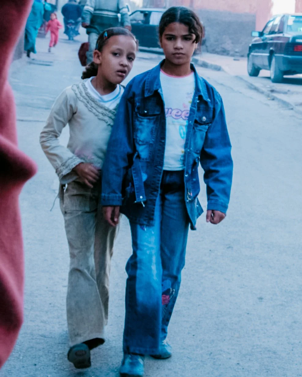 Twee jonge meisjes staan samen op een straat in een stad, met auto's op de achtergrond en anderen die voorbij lopen.