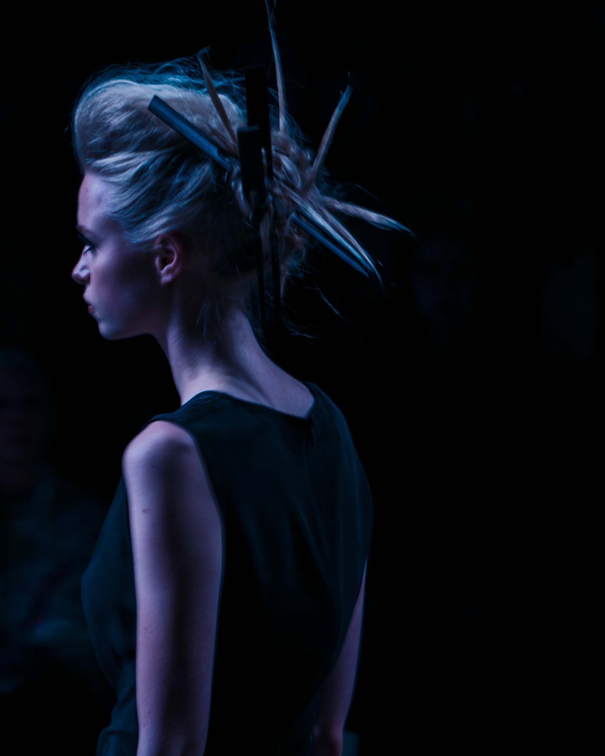 Een vrouw met blond haar en een zwarte jurk, haar haar is gestyled met grote speelse pennen en scharen. Ze staat zijwaarts afgebeeld tegen een donkere achtergrond.