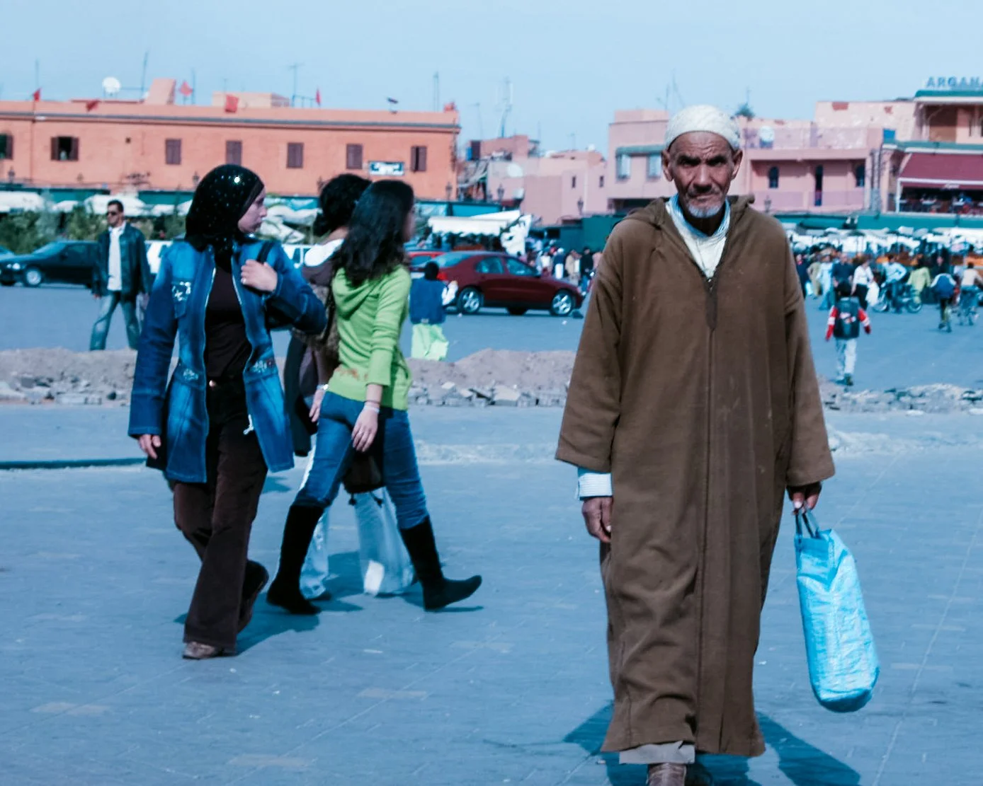 Man in een lange bruine jurk en wit hoofddoek, wandelend op een marktplein, met mensen op de achtergrond.
