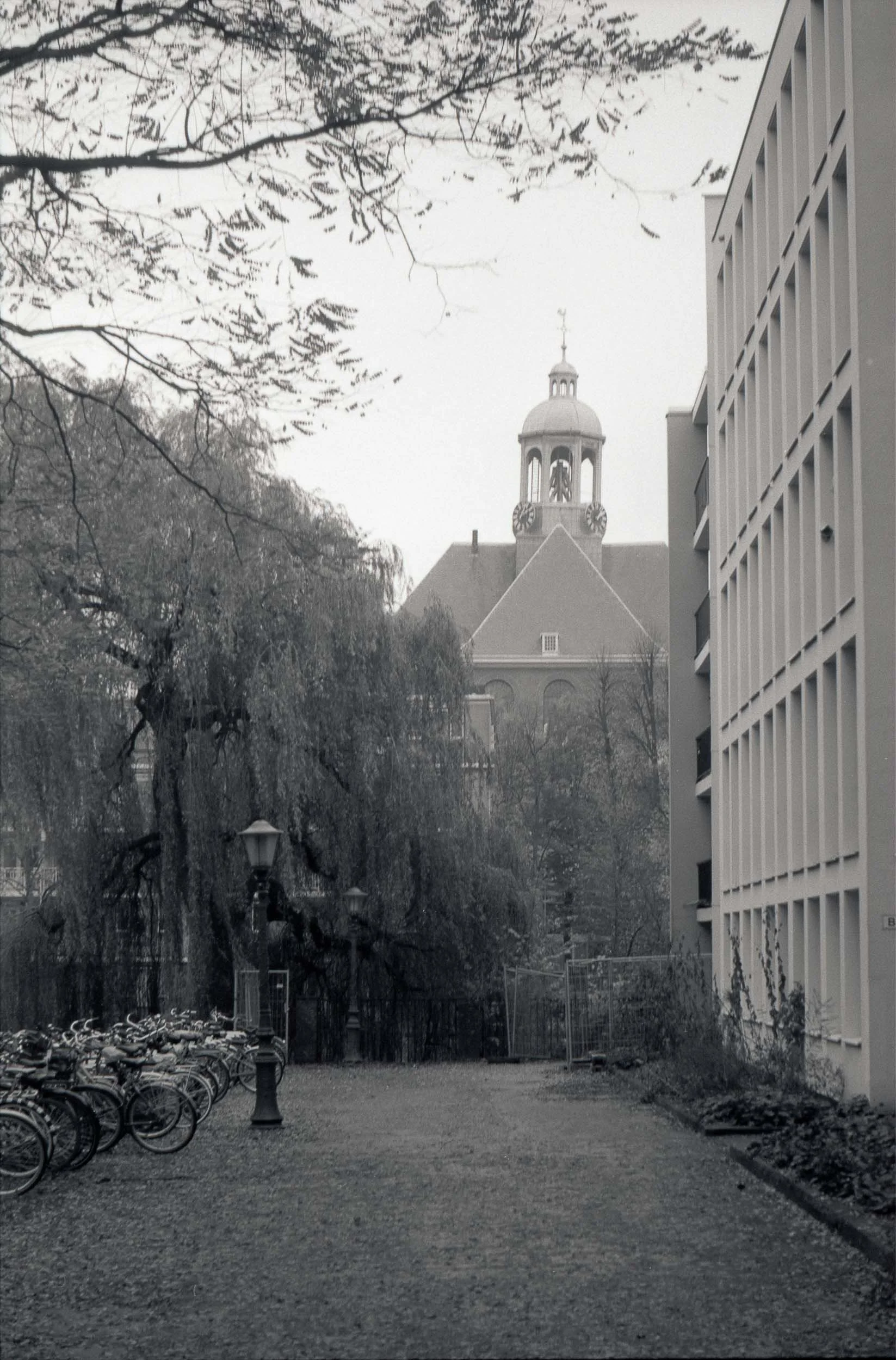 Een straat met fietsen links, bomen en een historisch kerkje op de achtergrond, onder een grijze lucht, in zwart-wit.