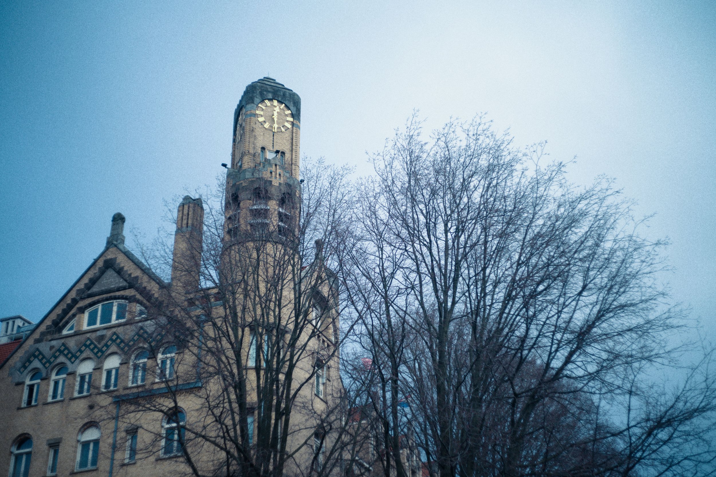 Een oude toren met een wijzerplaat en een klok, tussen blarenloze bomen, onder een grijze lucht.