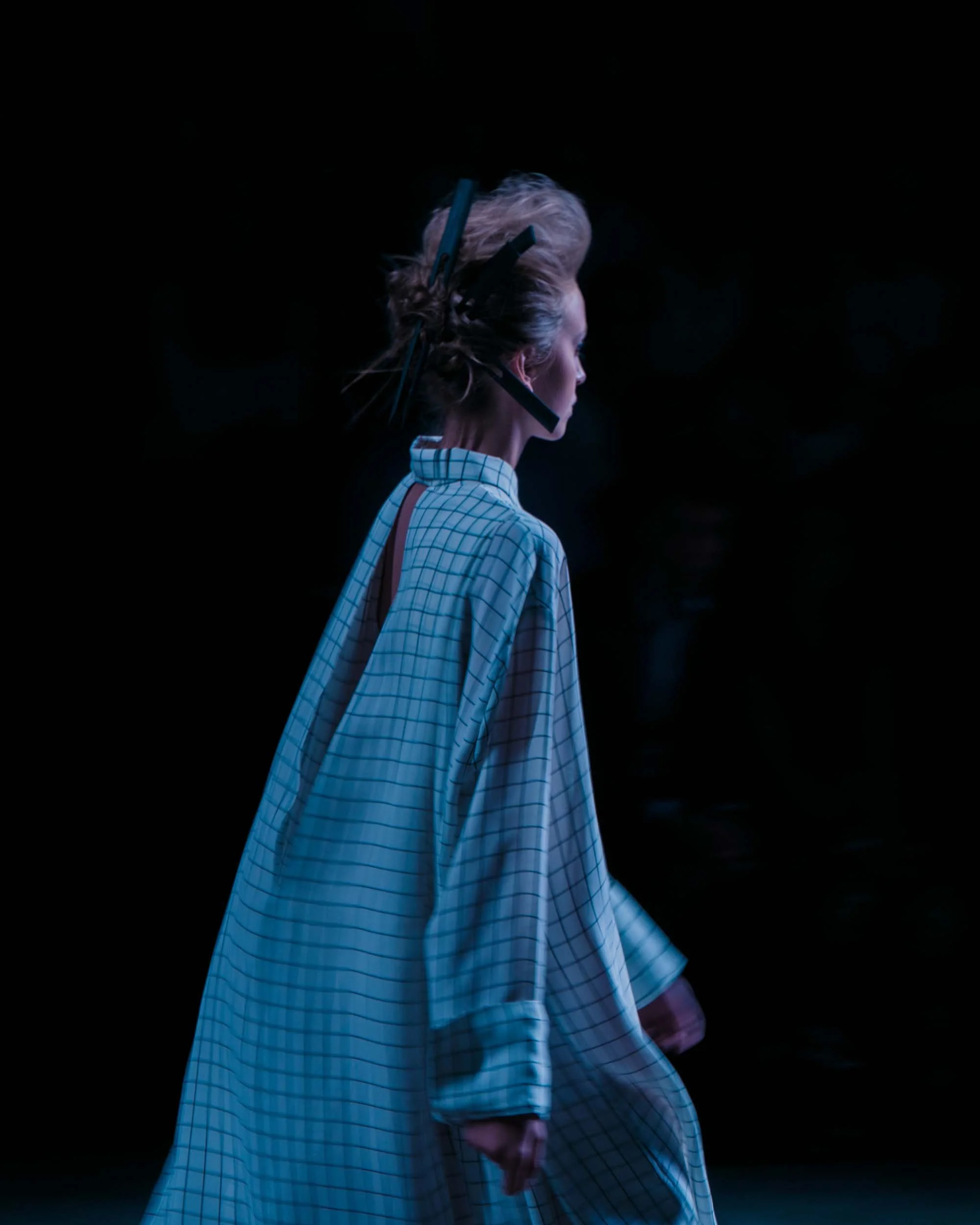 Een vrouw in een grote, getailleerde blouse met een geruite patroon, zichtbaar van achteren, met haar haar in een niet-voorzichtige knot en een hoofdband, staand in een donkere omgeving.