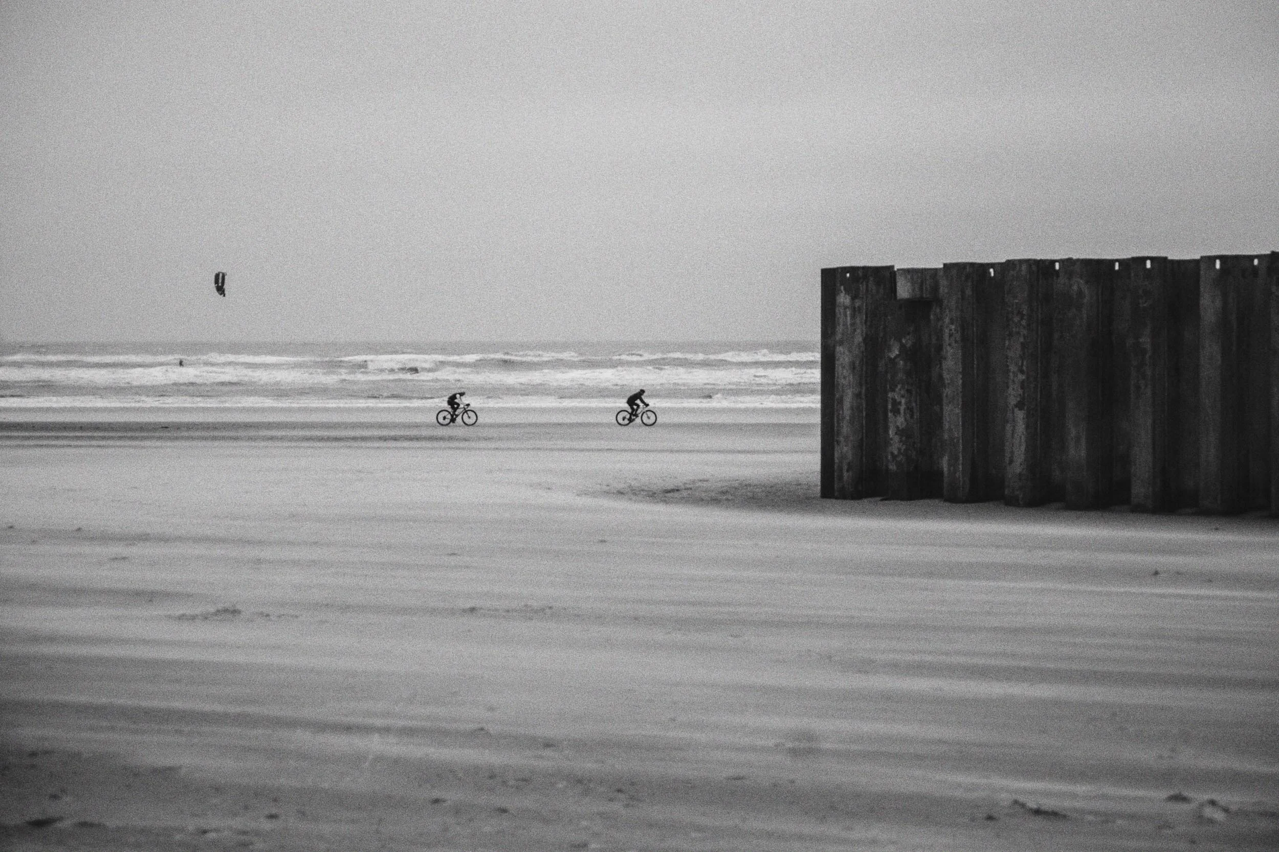 Zwart-wit foto van een lege strand met twee fietsers in de verte en een grote, oude zinken muur aan de rechterkant, met de oceaan en een kite surfer in de achtergrond.