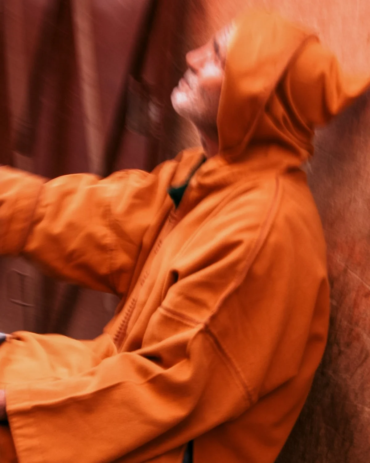 Een persoon met een baard, gekleed in een oranje jas met de capuchon op, die zich naast een muur of deur bevindt.