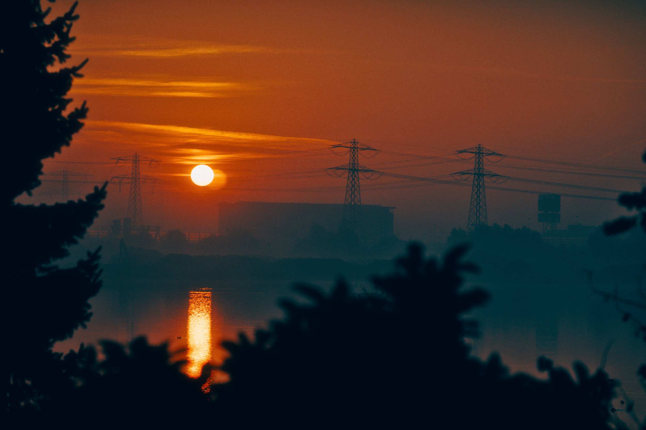 De zon onder de horizon met een reflectie op het water, omgeven door silhouetten van bomen en hoogspanningsmasten in de afstand.