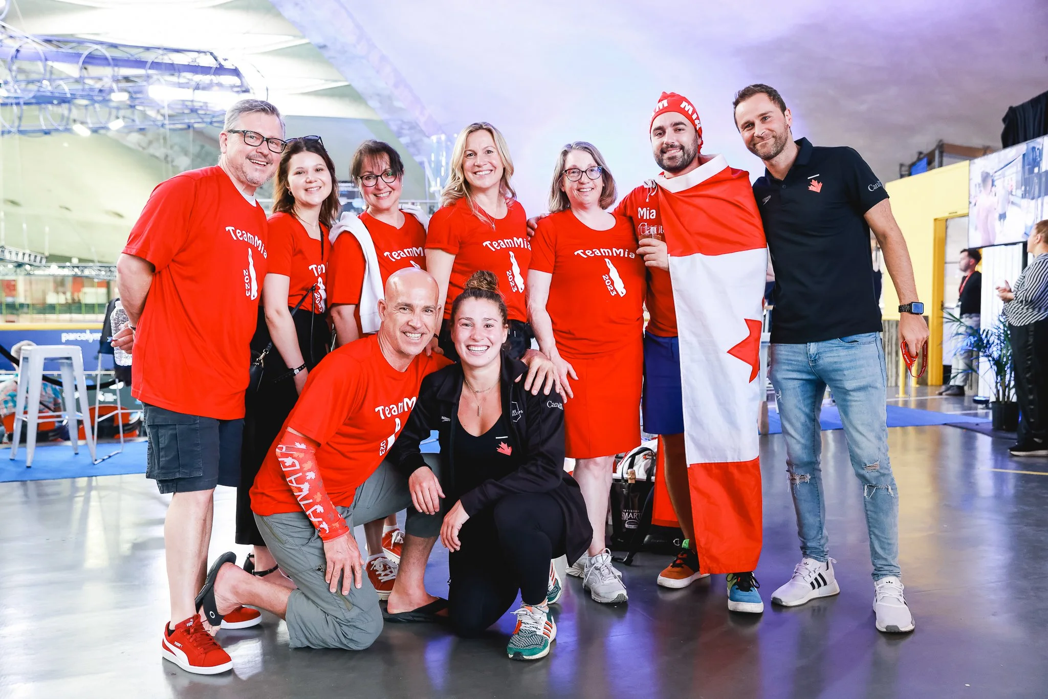 Groupe de personnes souriantes portant des t-shirts rouge avec l'inscription 'TeamMia' à l'intérieur d'un stade ou centre sportif.