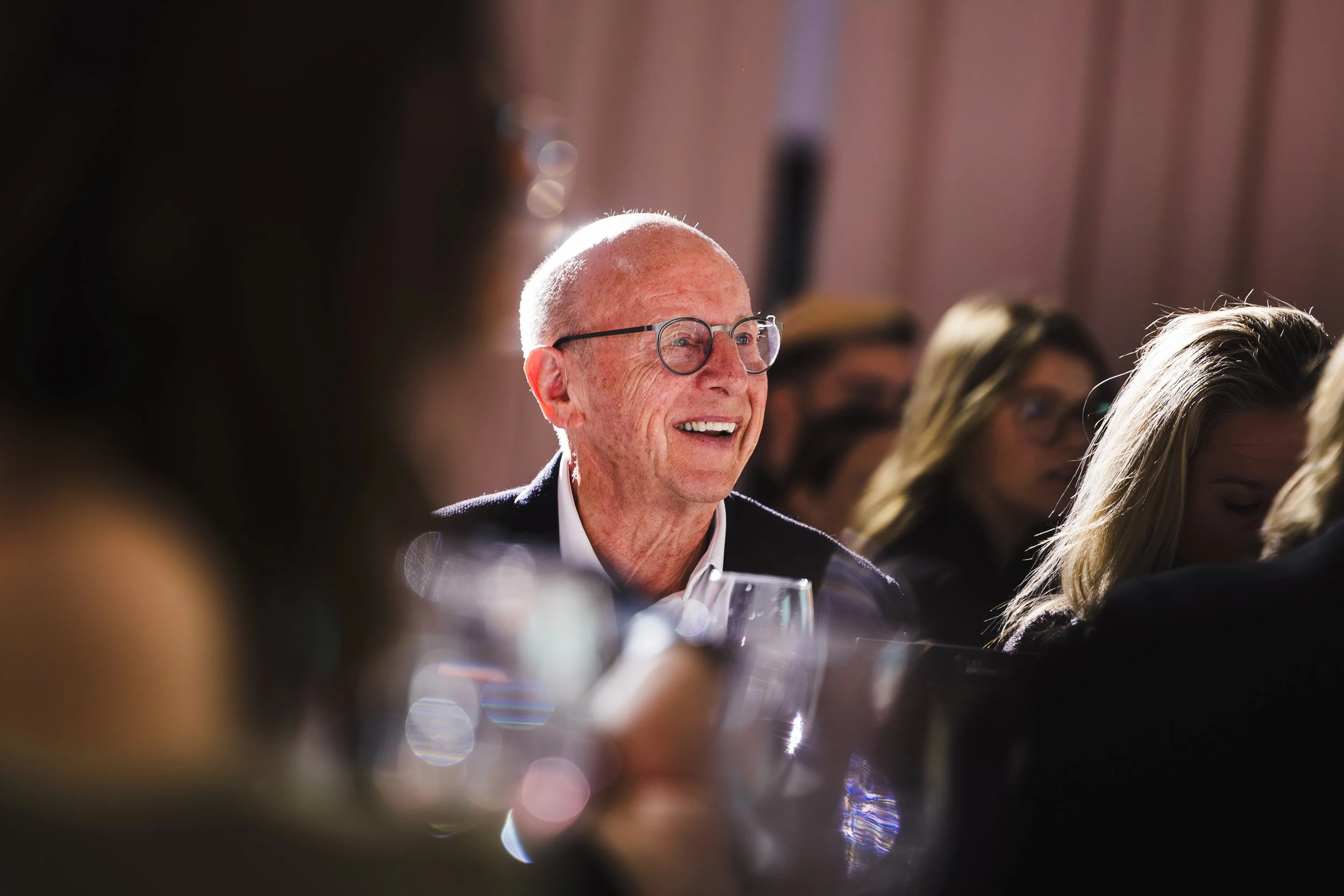 Un homme âgé avec des lunettes, souriant lors d'un repas ou d'une événement social, entouré de plusieurs personnes lors d'une réunion ou d'une célébration.