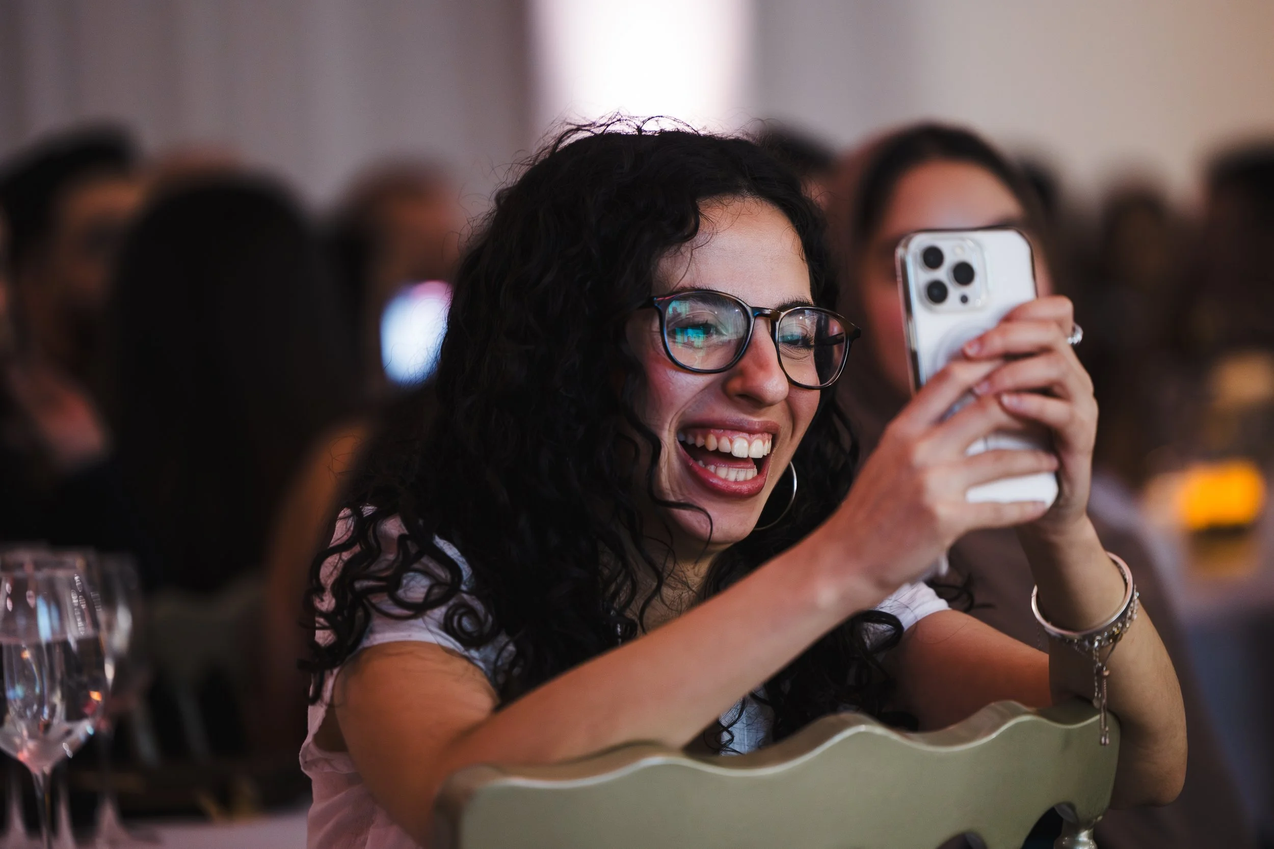 Une femme souriante et portant des lunettes tient un téléphone portable pour prendre une photo ou enregistrer une vidéo, dans une salle avec un groupe de personnes en arrière-plan.