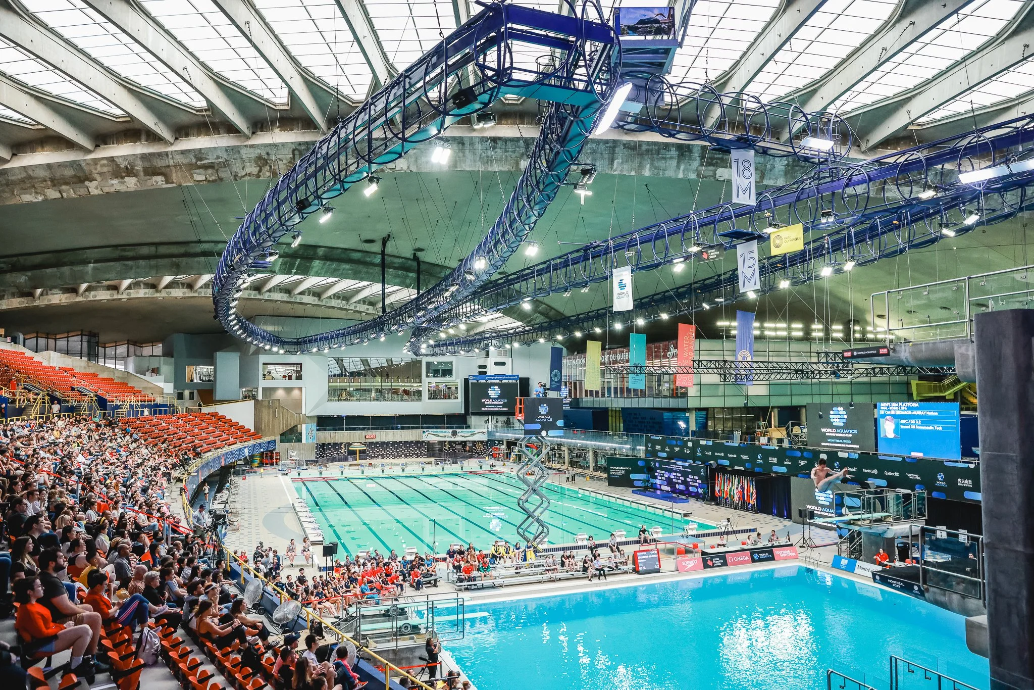 Piscine olympique intérieure avec spectateurs assis sur les gradins, dômes en verre au plafond, décorations et affichages liés à la plongée et à la natation.