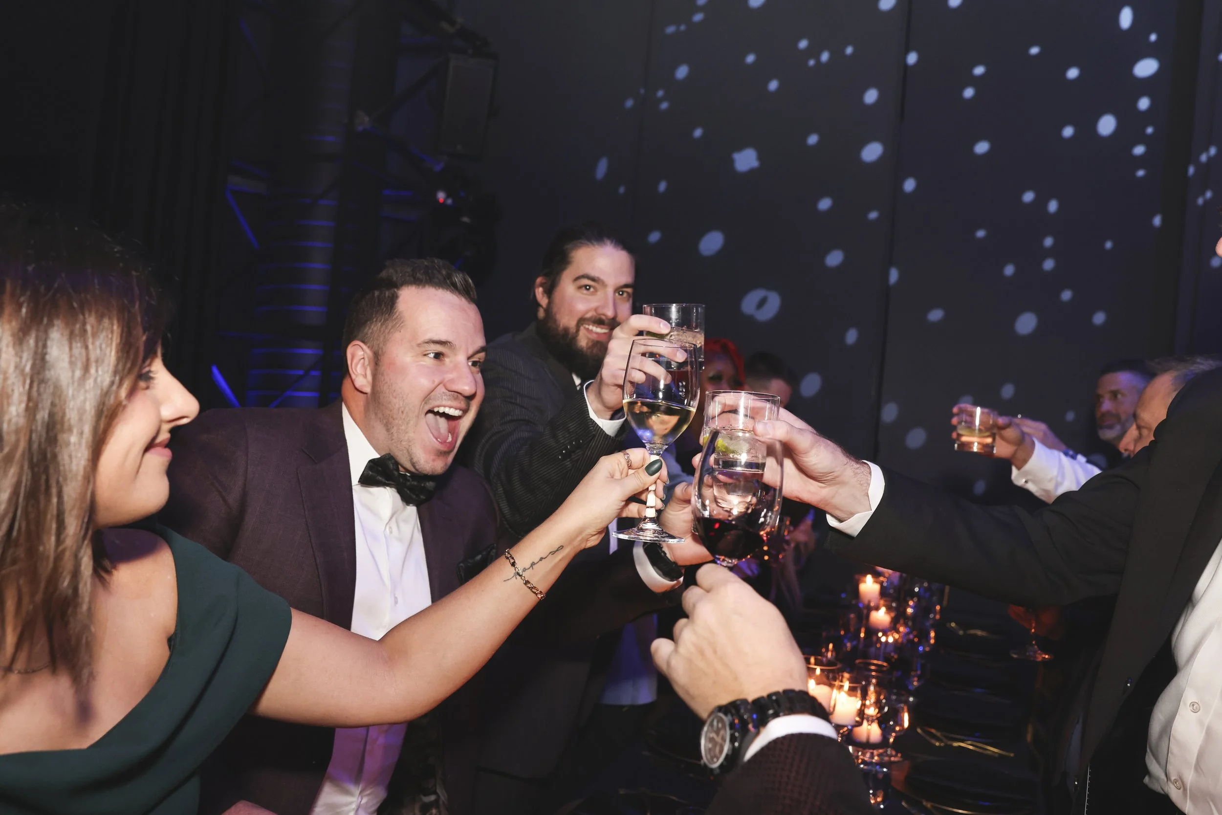 Groupe de personnes habillées élégamment faisant un toast lors d'une célébration dans un décor romantique avec bougies et projection de lumières sur le mur.