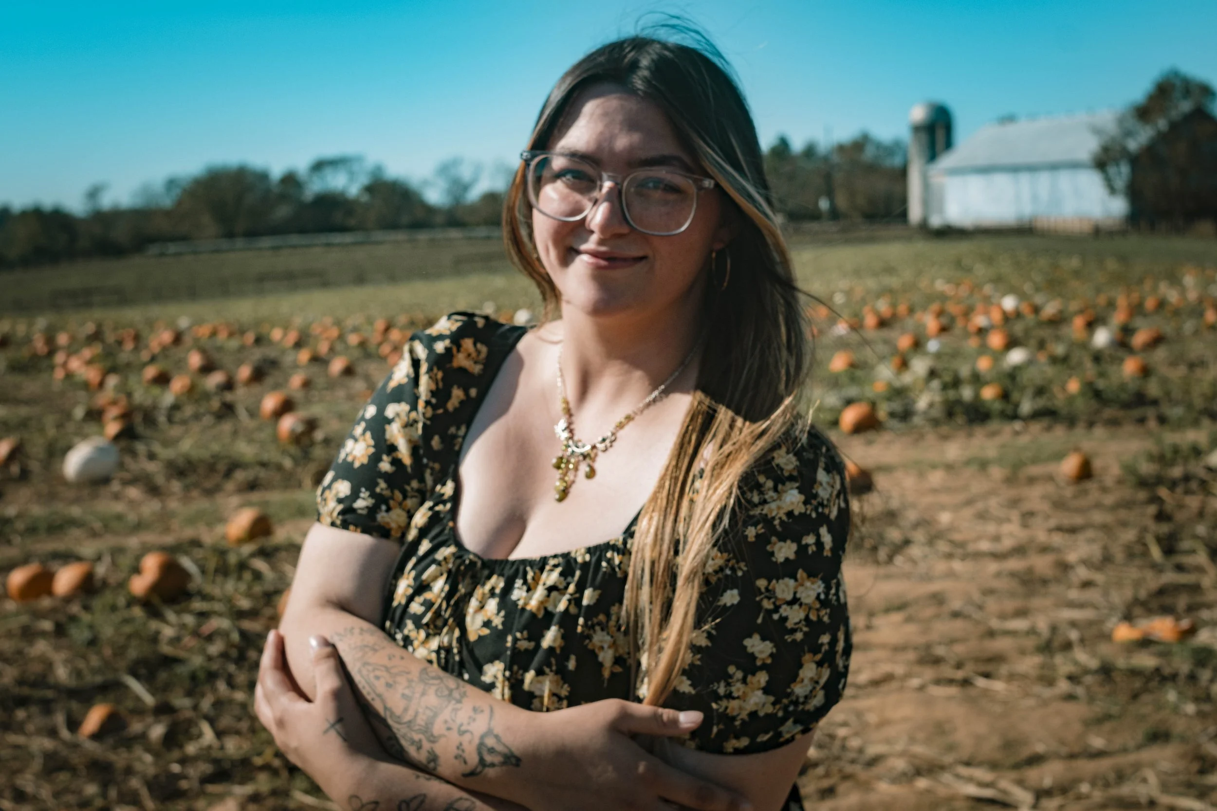 A young woman with long hair, glasses, and tattoos on her arms stands in a pumpkin patch on a sunny day, smiling at the camera.