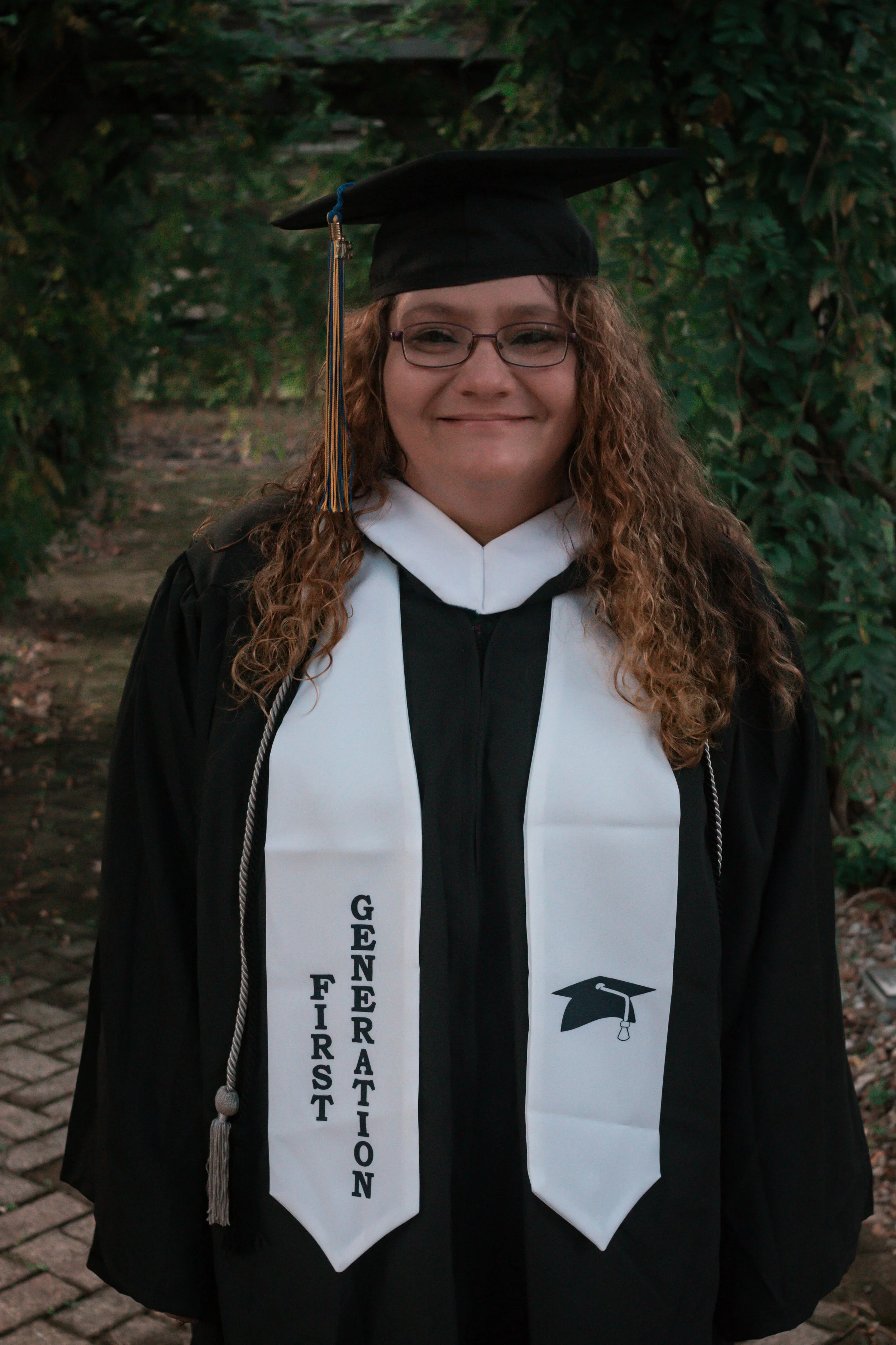 A woman in a black graduation cap and gown, wearing a white stole with black text and a graduation cap symbol, outdoors in front of green foliage.