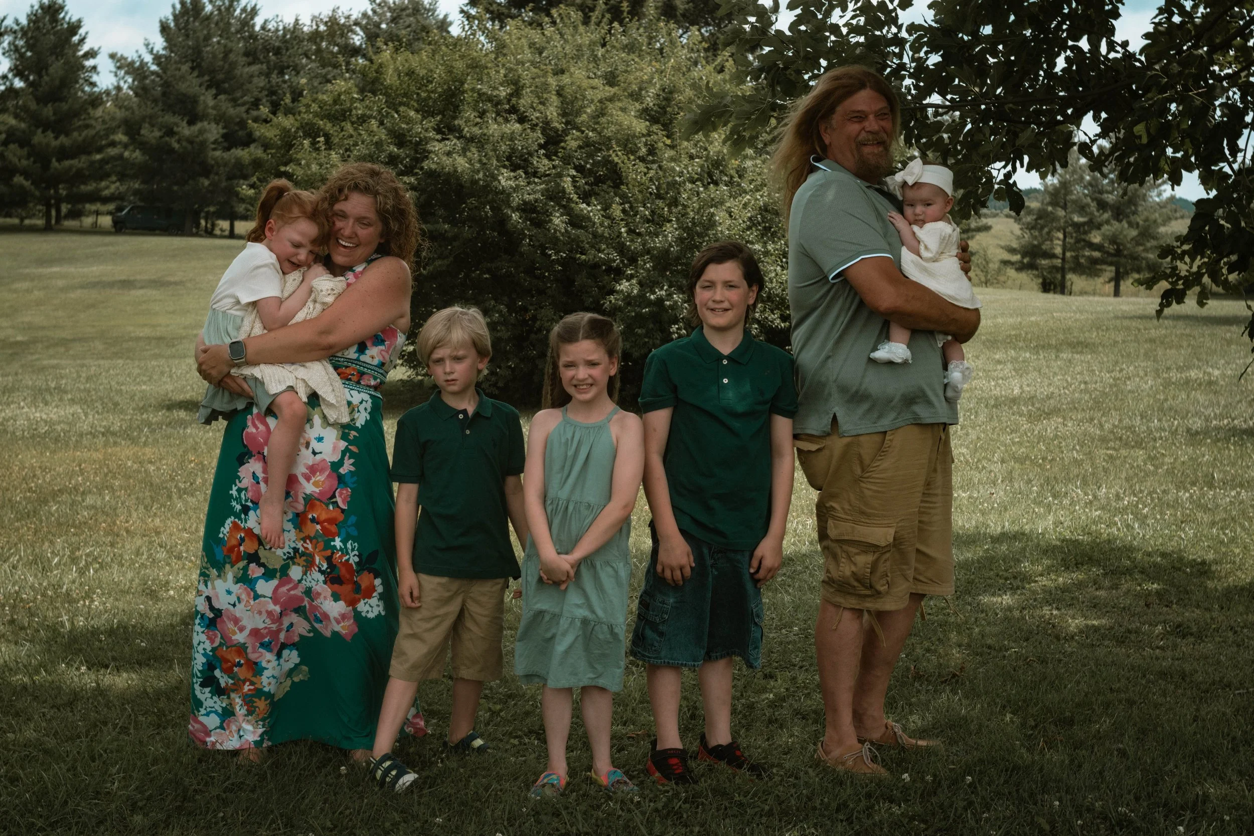 Family photo of two adults and four children standing outdoors on a grassy field with trees in the background.