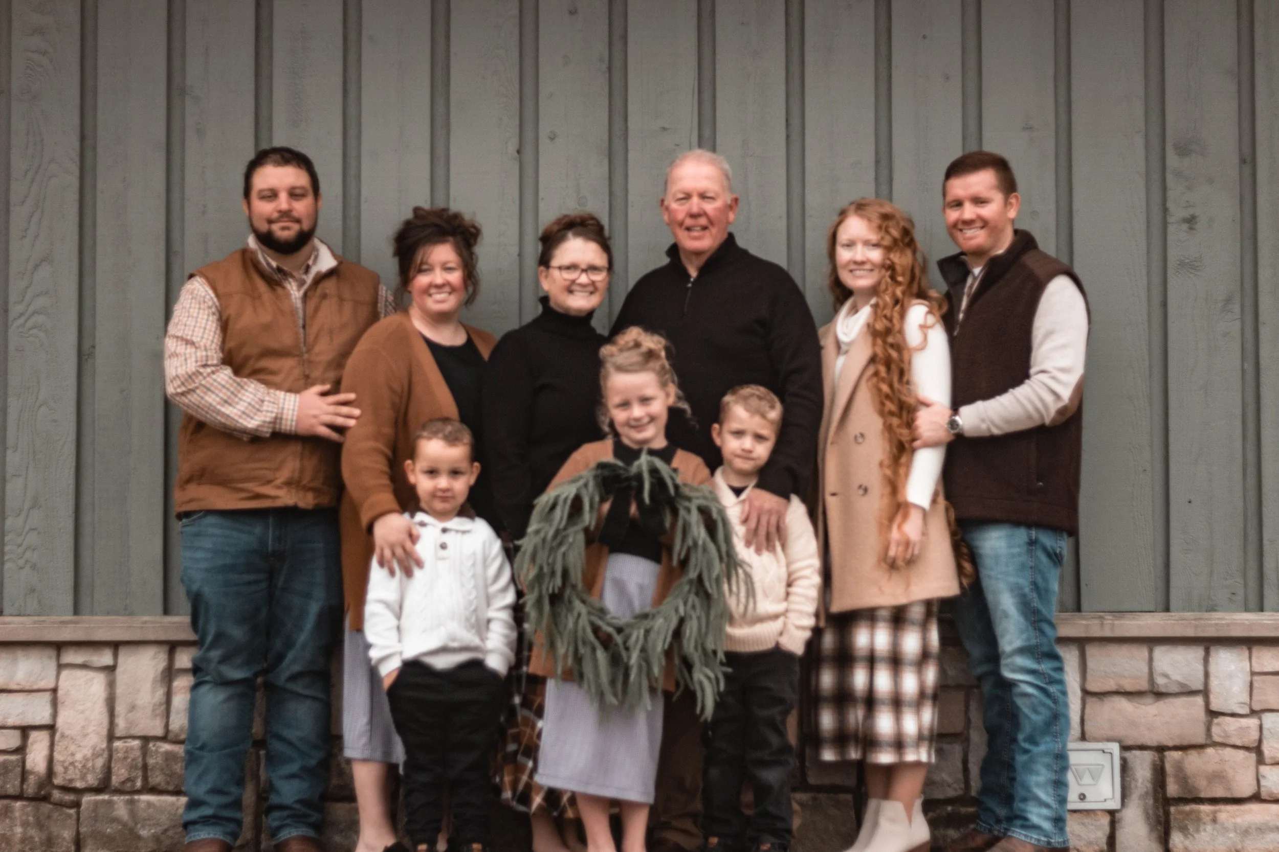 Family of ten, including three children, standing outside in front of a green wooden wall, posing for a group photo.