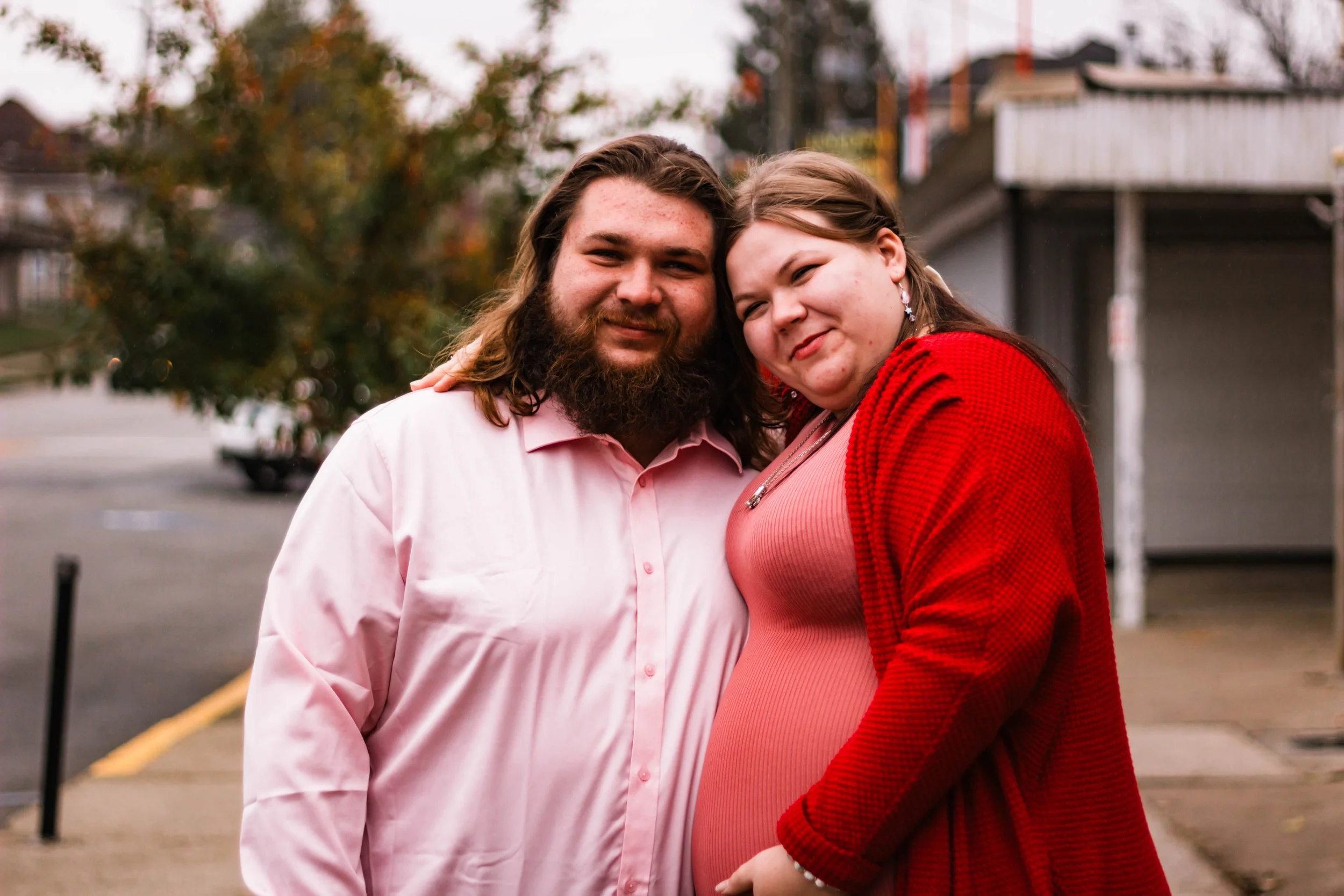 A couple standing close together outdoors, with the woman appearing pregnant, smiling at the camera during daytime.