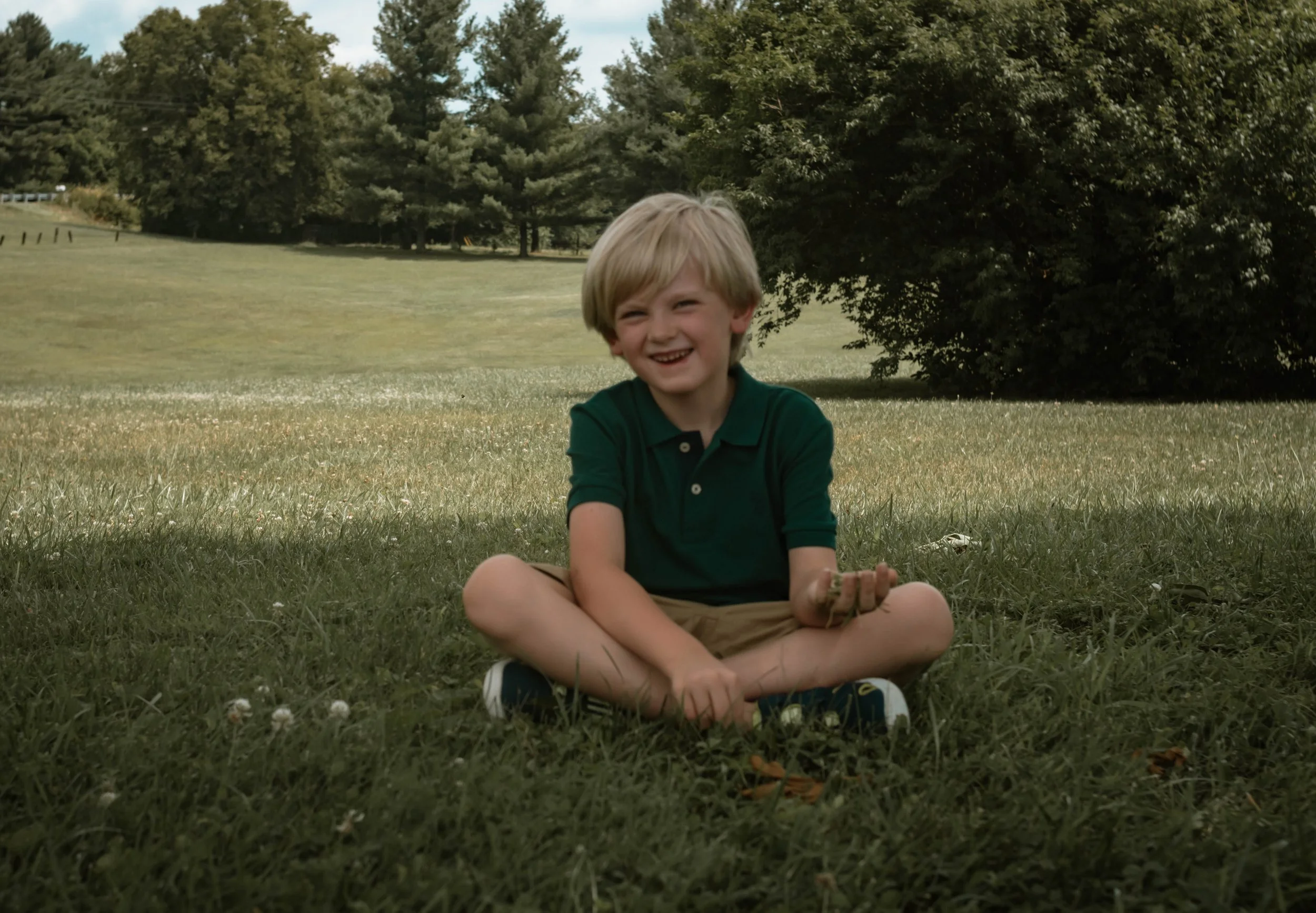A young boy with blonde hair, sitting cross-legged on grass in a park, smiling, wearing a dark green polo shirt and khaki shorts.