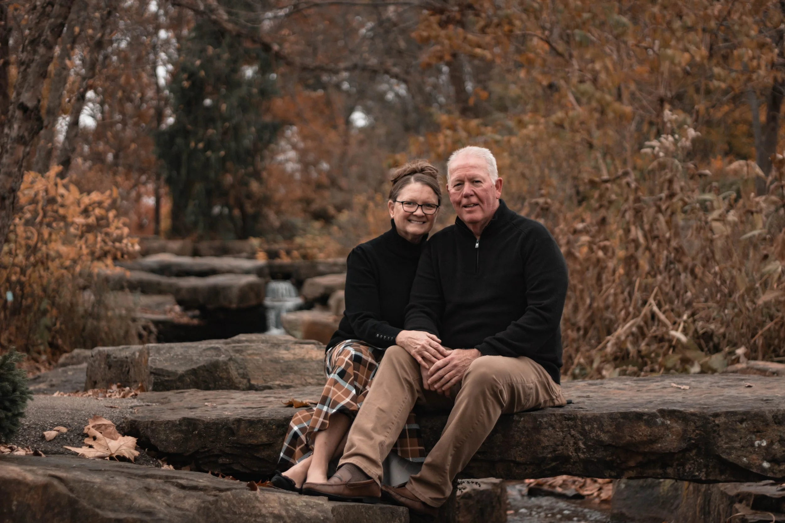 An elderly couple sitting on a large rock in a park with fall foliage in the background, smiling at the camera.
