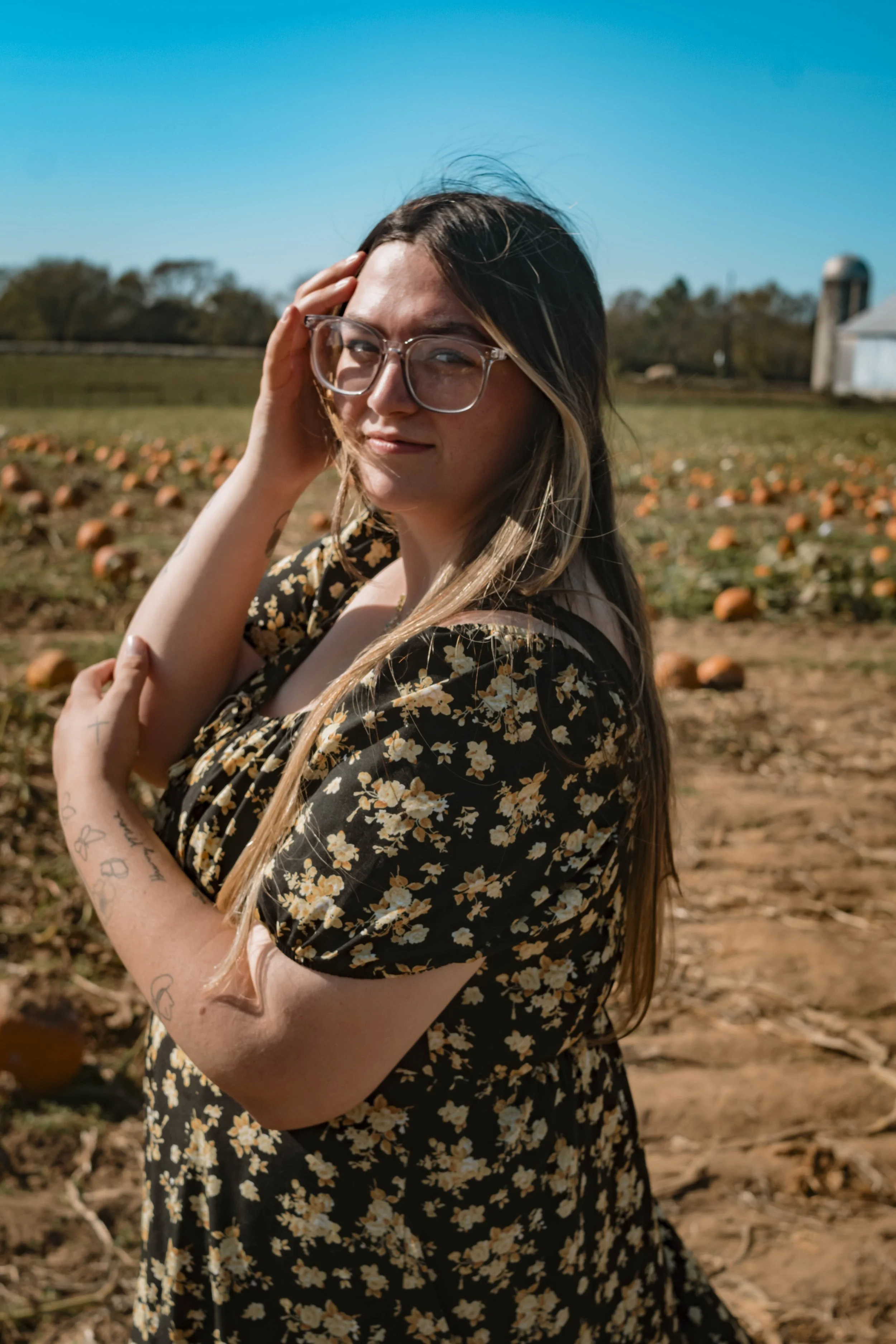 A woman with long hair and glasses standing in a pumpkin patch on a sunny day, wearing a black floral dress.