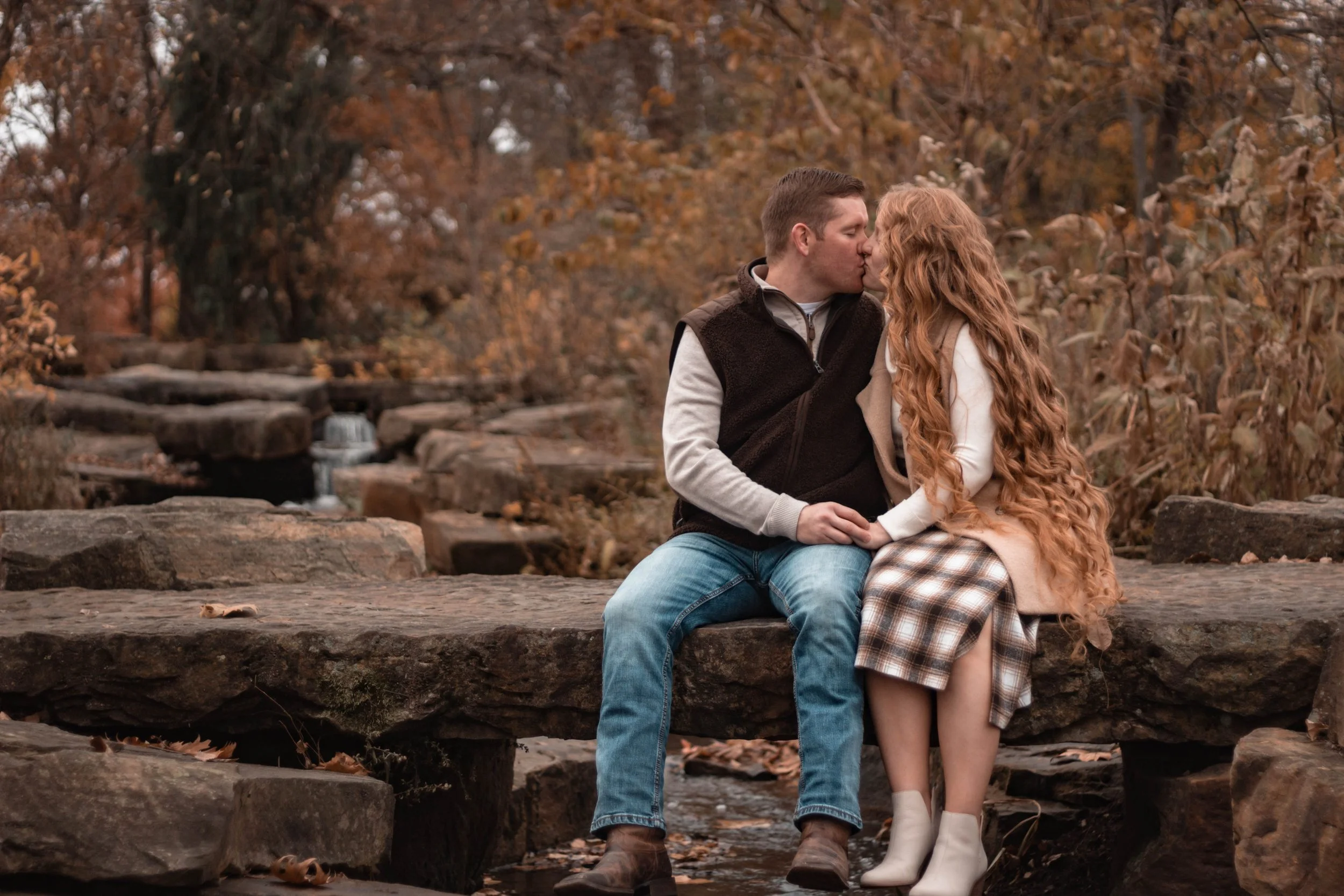 A couple sharing a kiss while sitting on a stone bench in a forest with fall foliage.
