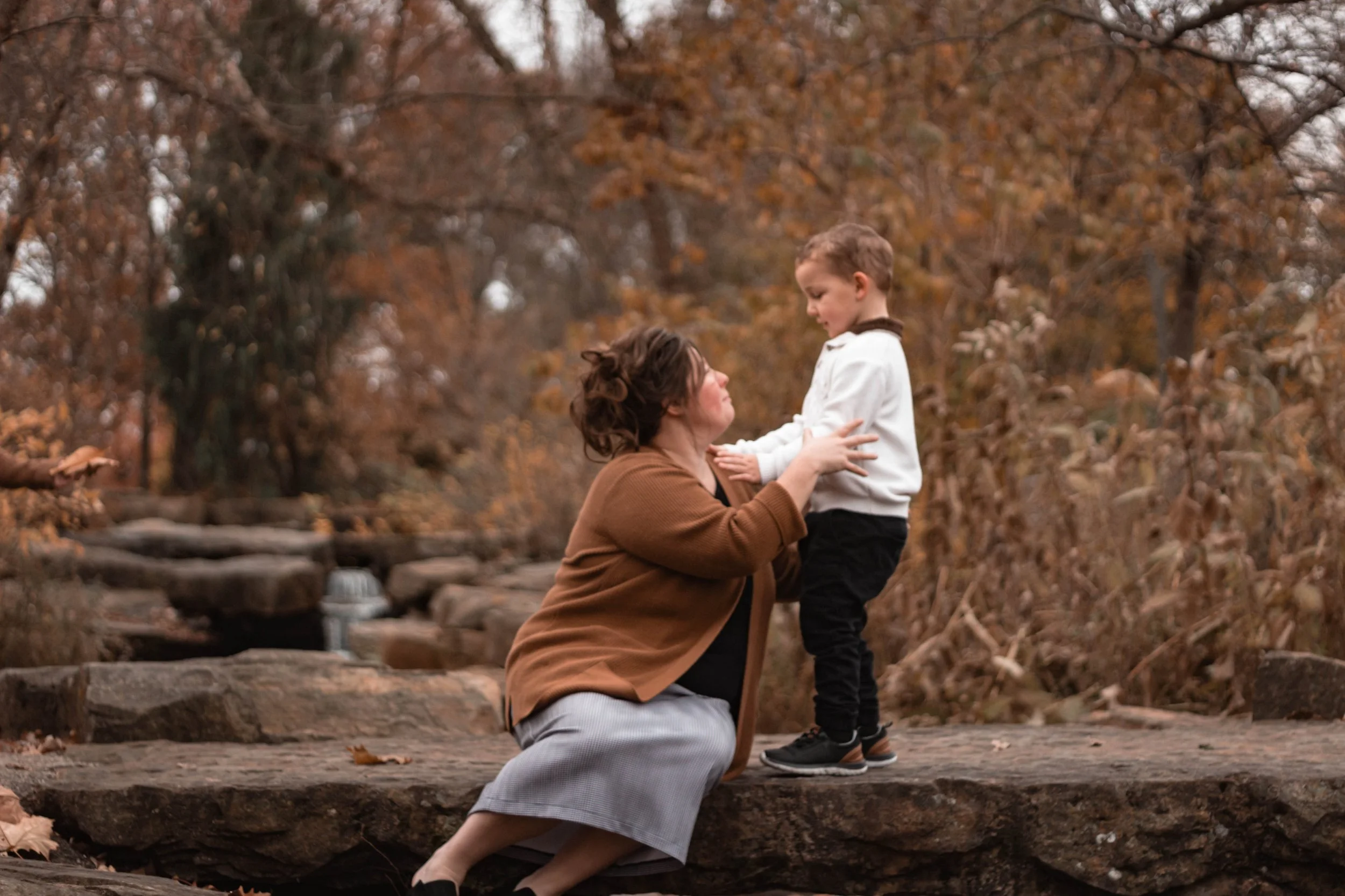 A woman kneeling on a large rock, holding hands with a young boy on a forest trail during fall, surrounded by autumn leaves and trees.