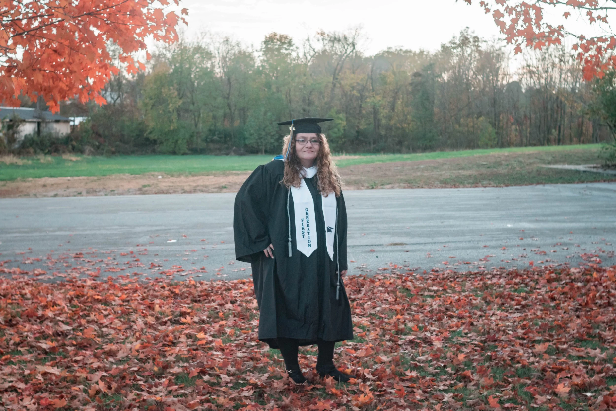 A woman in a graduation cap and gown standing outdoors on a bed of fallen autumn leaves, with trees and a paved area in the background.