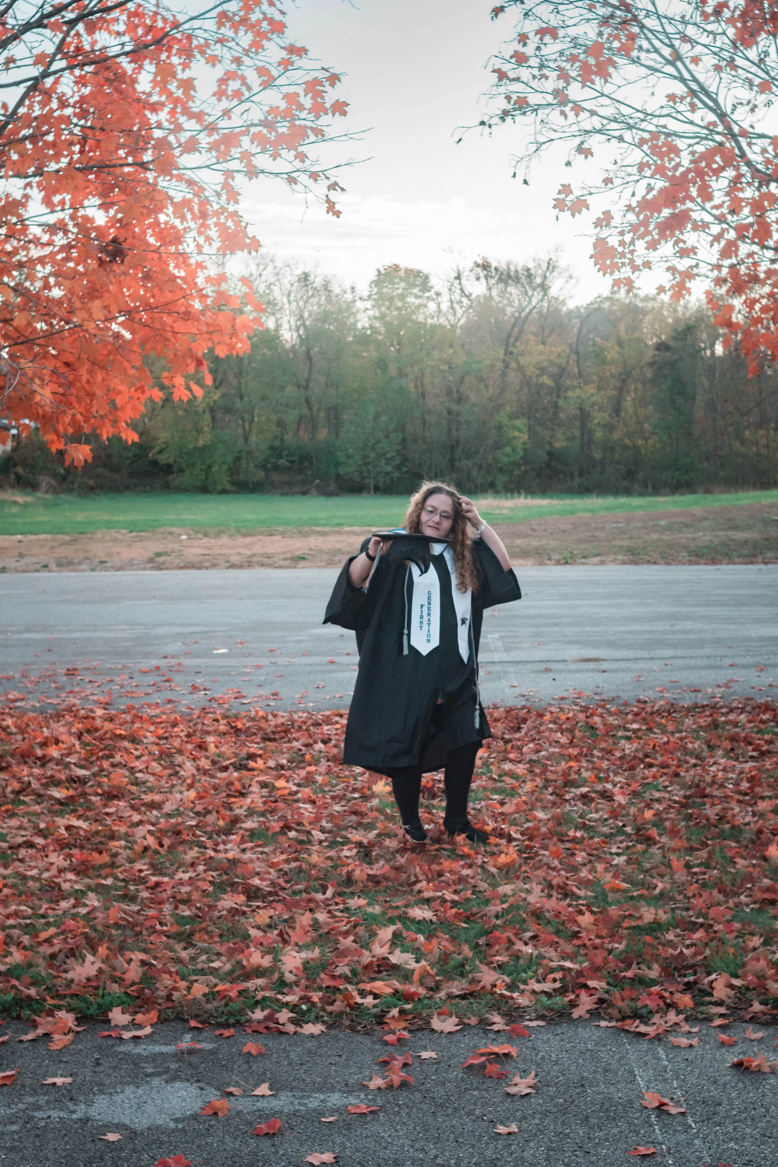 A girl in a graduation gown and cap standing outdoors among fallen autumn leaves, holding her cap in one hand and touching her hair with the other, with trees and a road in the background.