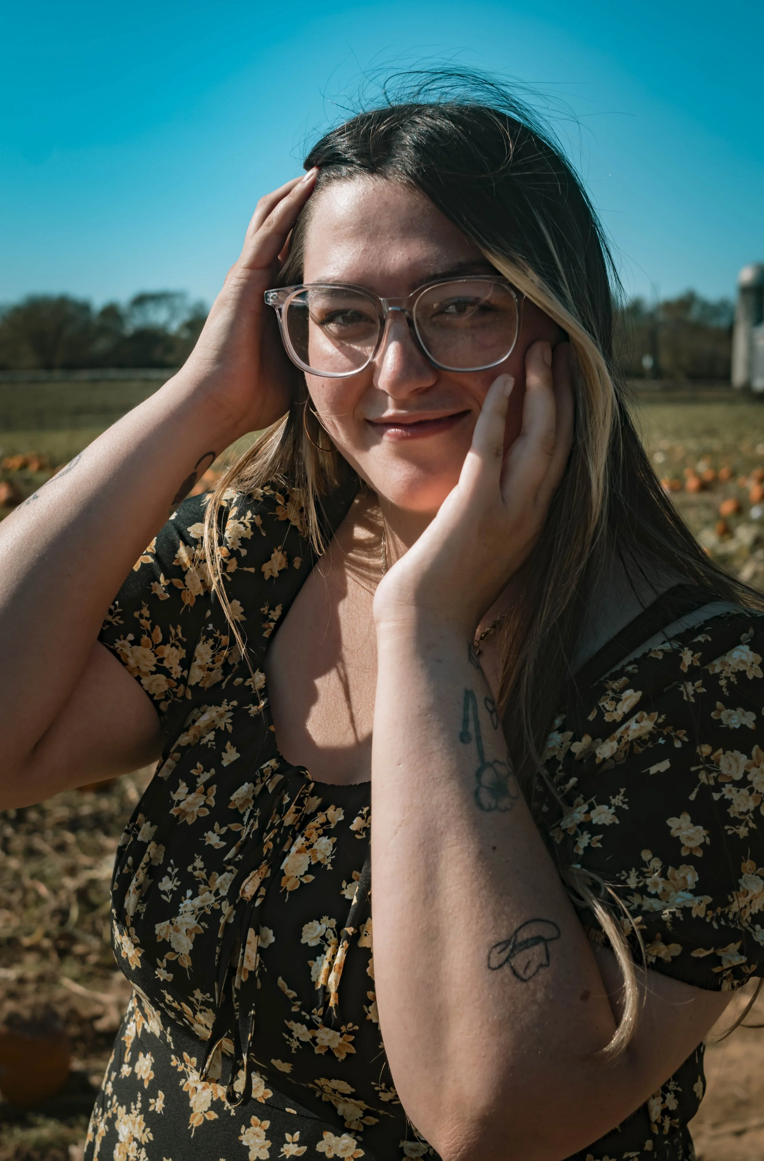 Young woman with glasses and tattoos outdoors on sunny day, smiling gently.