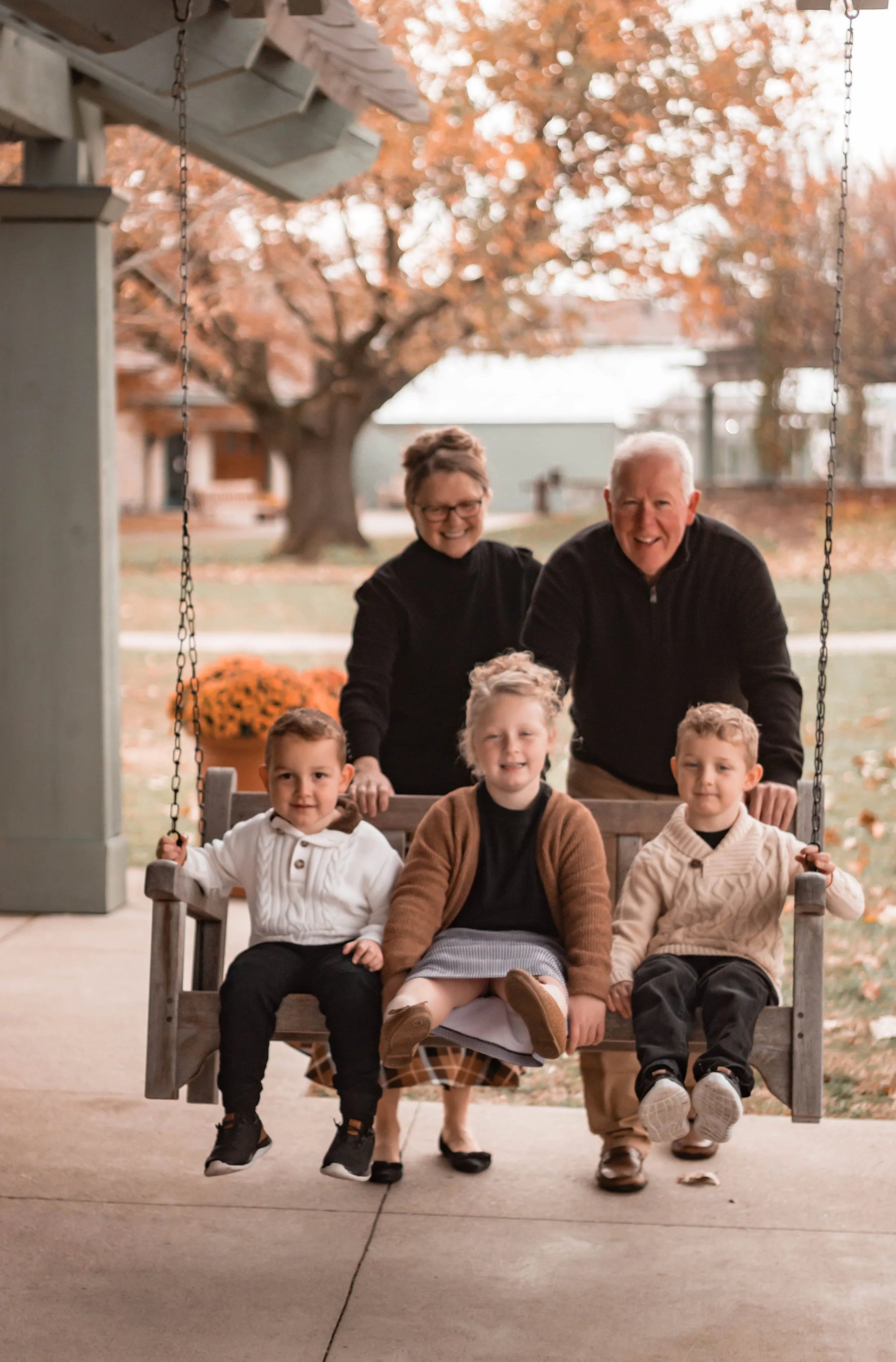 A family of six enjoying time on a porch swing during autumn. The group includes two older adults and four children, with some sitting on the swing and others standing behind it. Fallen leaves and a large tree with orange foliage are visible in the b