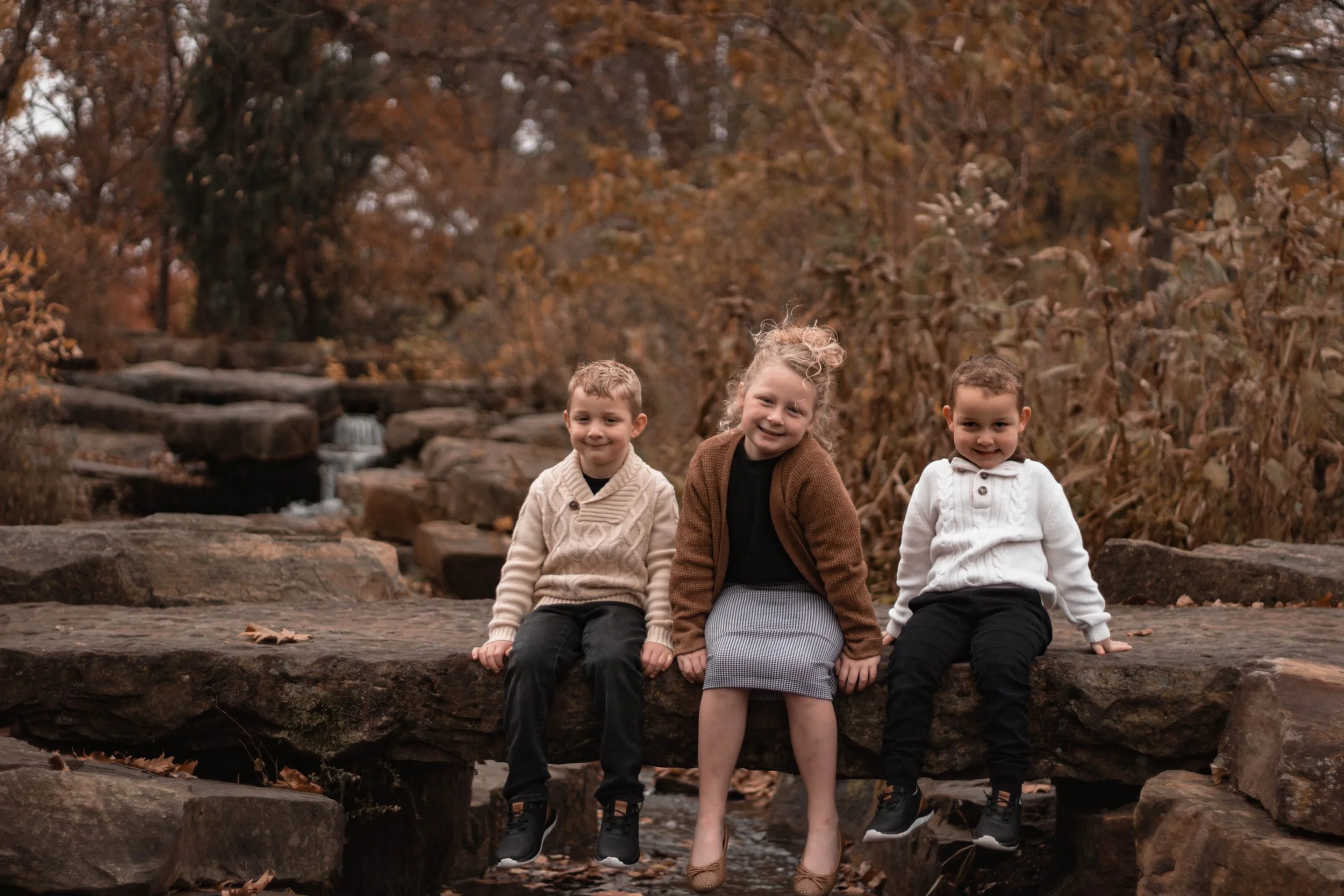 Three children sitting on a large rock in an autumn forest, smiling and posing for the camera.