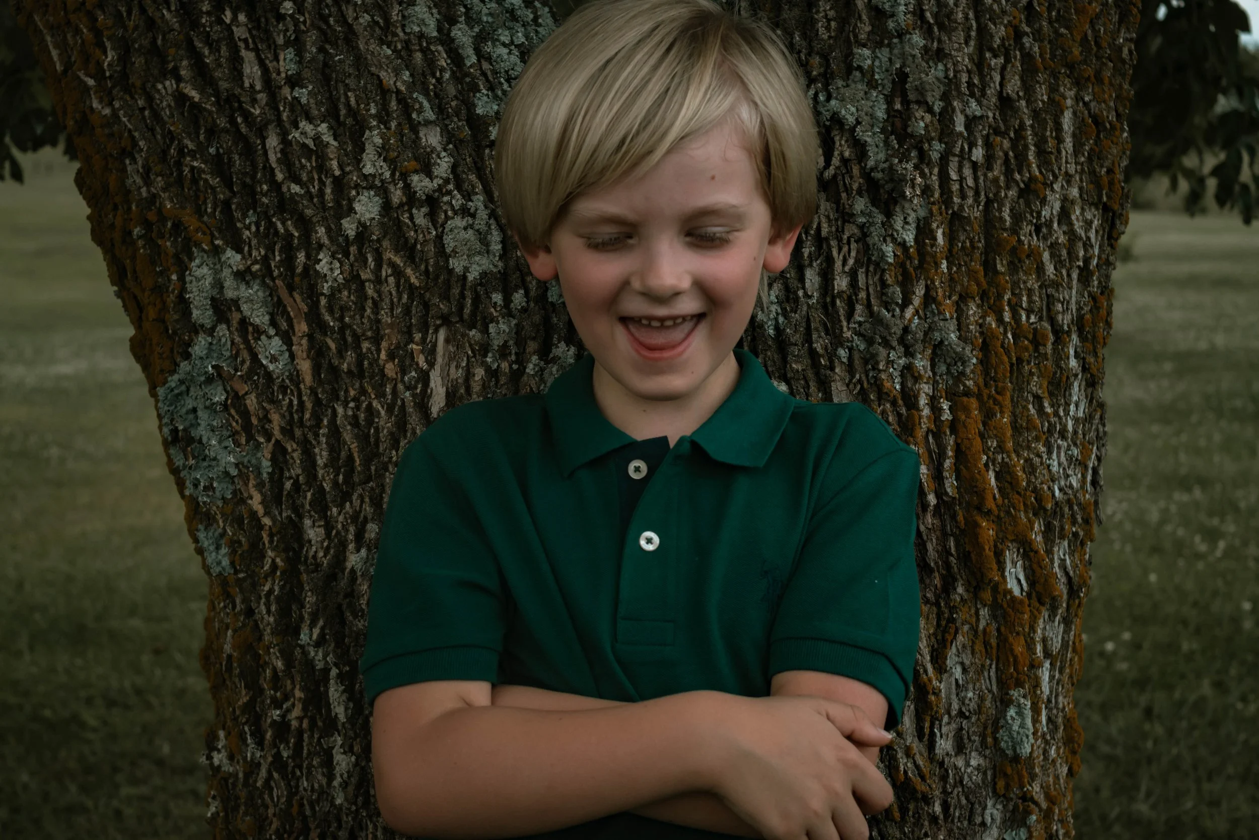 A young boy with blonde hair, wearing a green polo shirt, leaning against a tree with a broad smile and crossed arms, in an outdoor park setting.