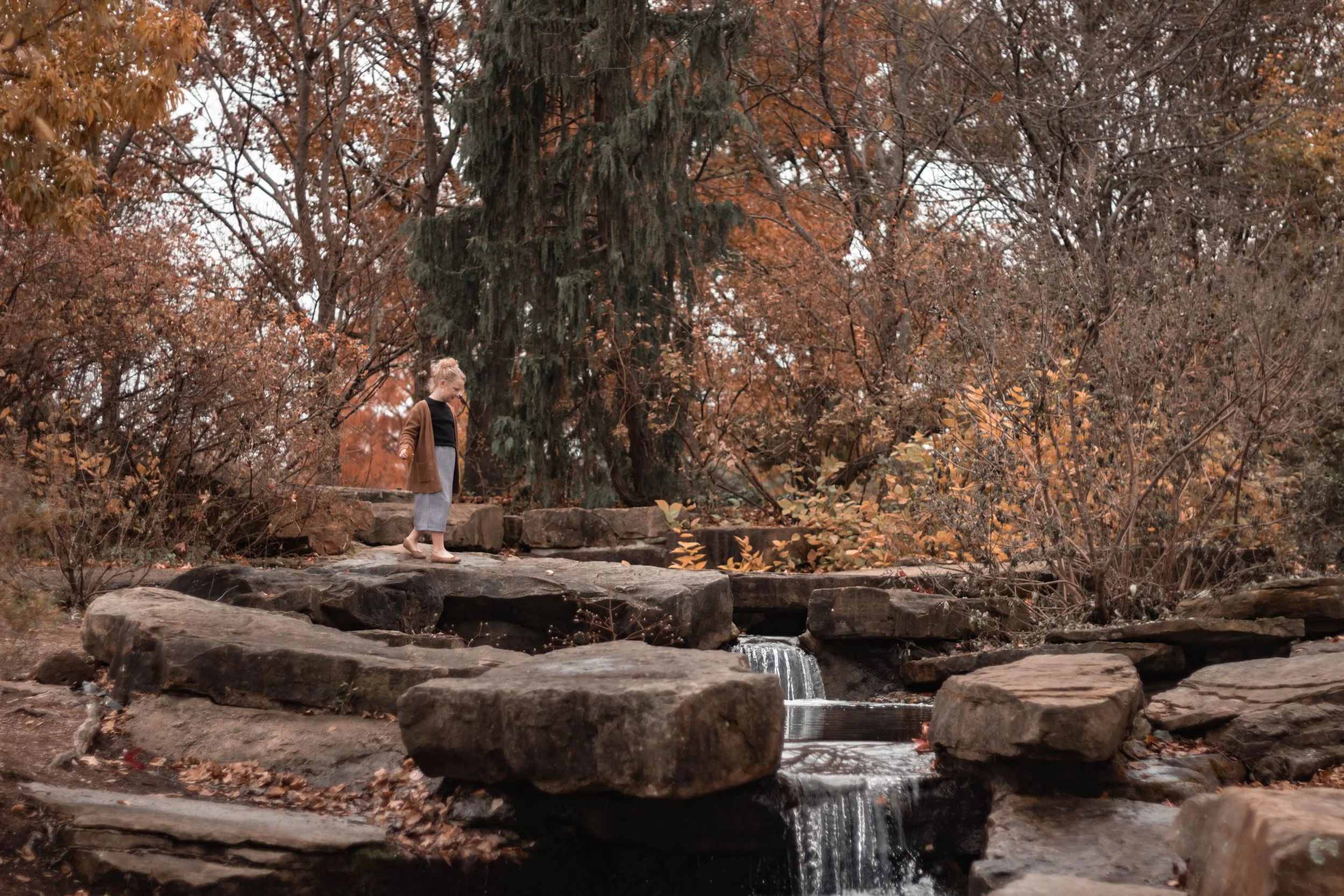A young girl with blonde hair in a bun, wearing a brown jacket, black top, and gray skirt, crossing a small rocky waterfall in a forest with autumn foliage.
