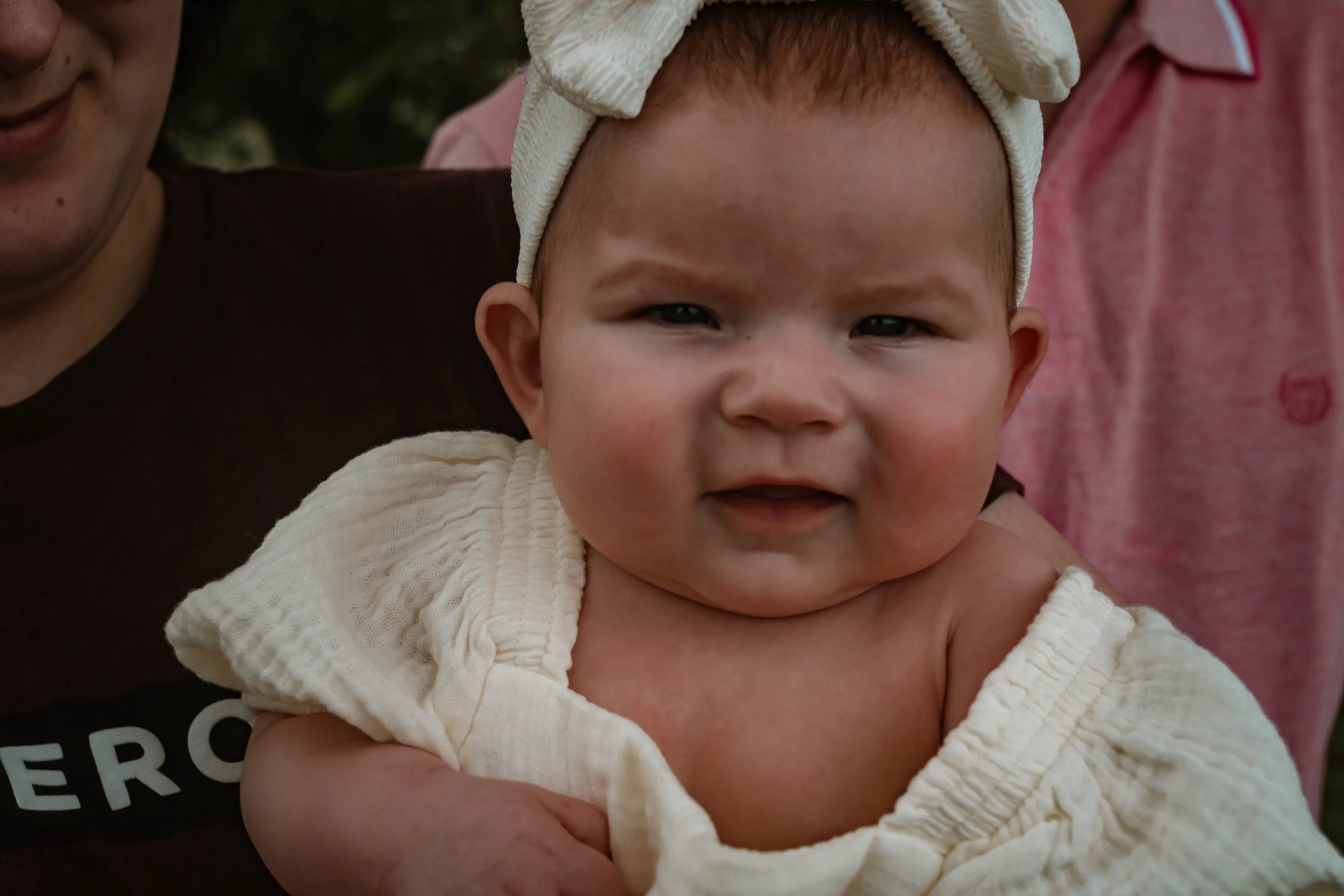 Close-up of a baby with red hair, wearing a cream-colored headband and a cream-colored towel draped over shoulders, held by adults.