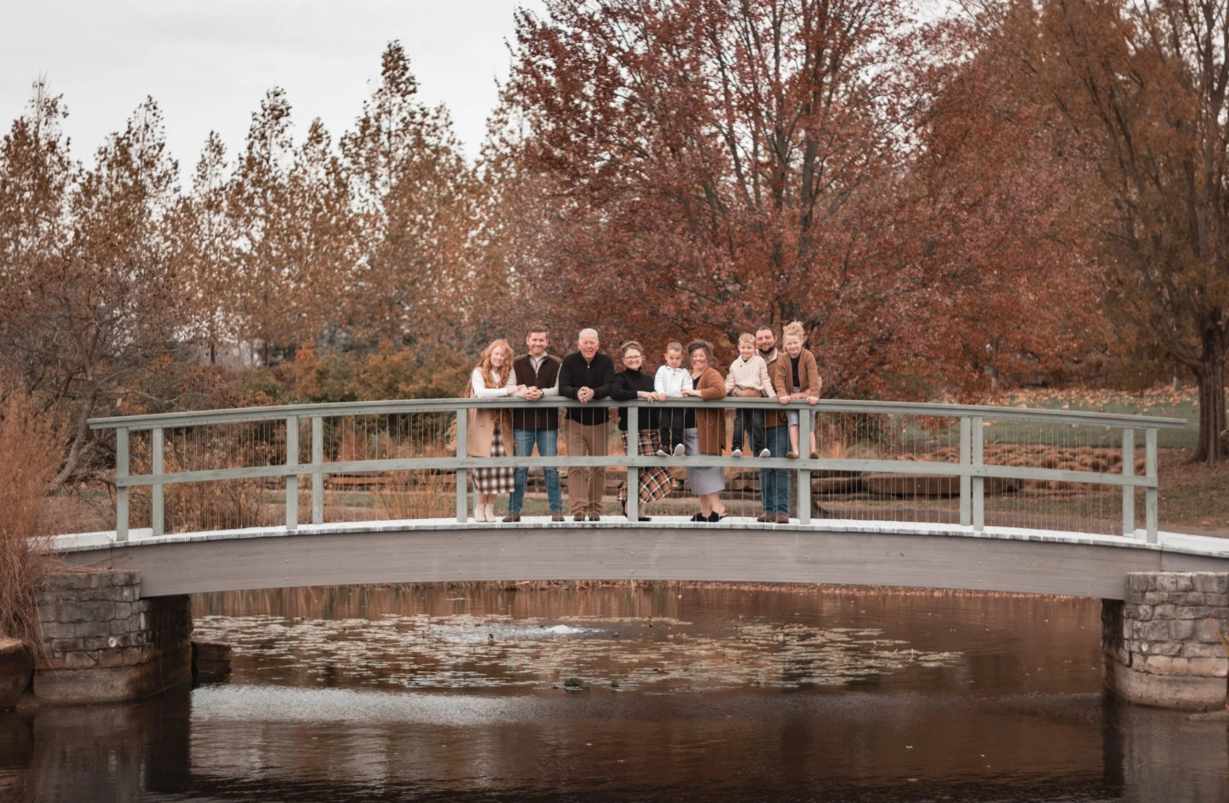 Family standing on a small bridge over a river, surrounded by autumn trees with orange and brown foliage.