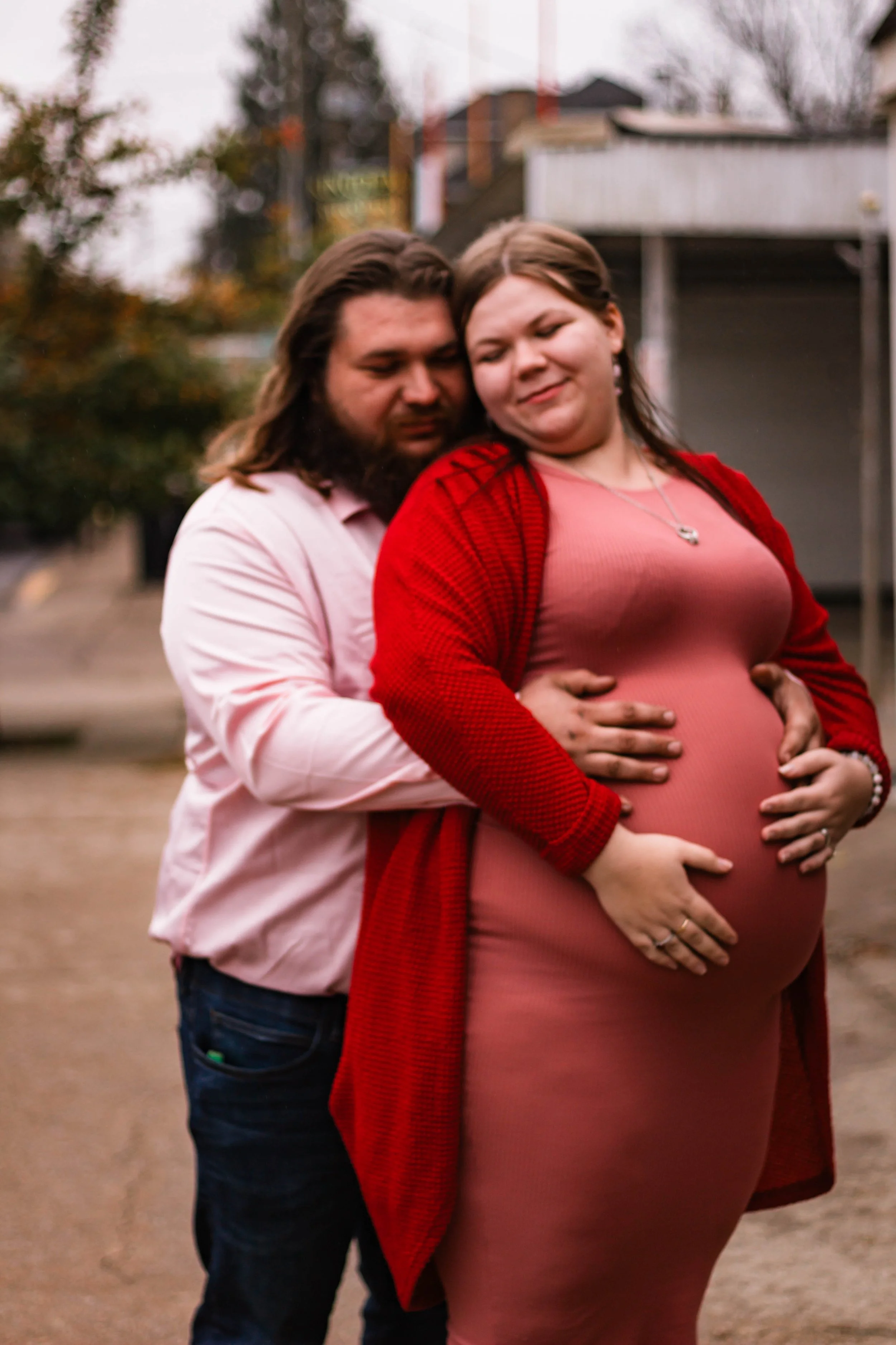 A pregnant woman in a pink dress and red cardigan holding her belly, with a man standing behind her, holding her and her belly, outdoors during autumn.
