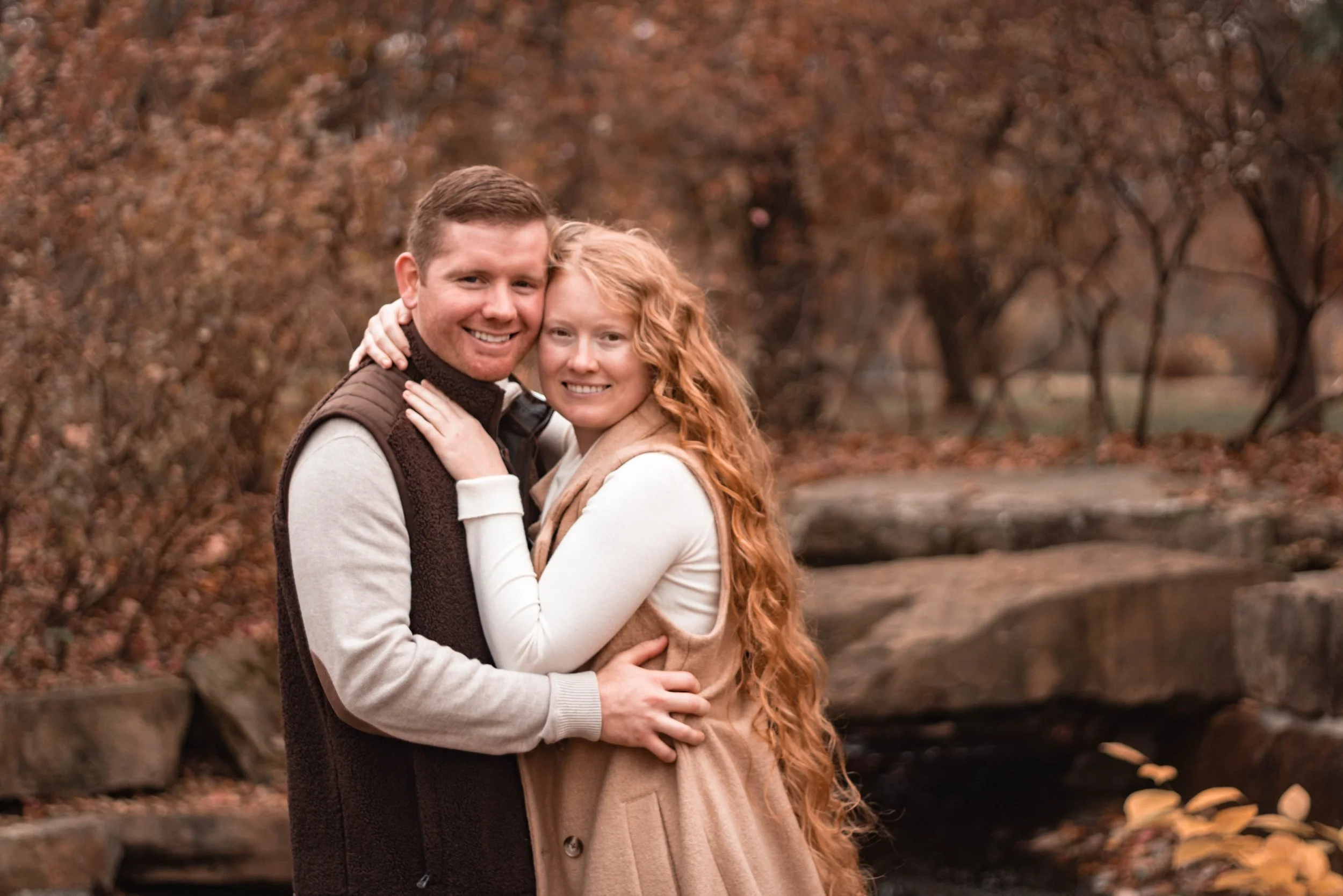 A happy couple hugging outdoors in autumn with fallen leaves and trees in the background.