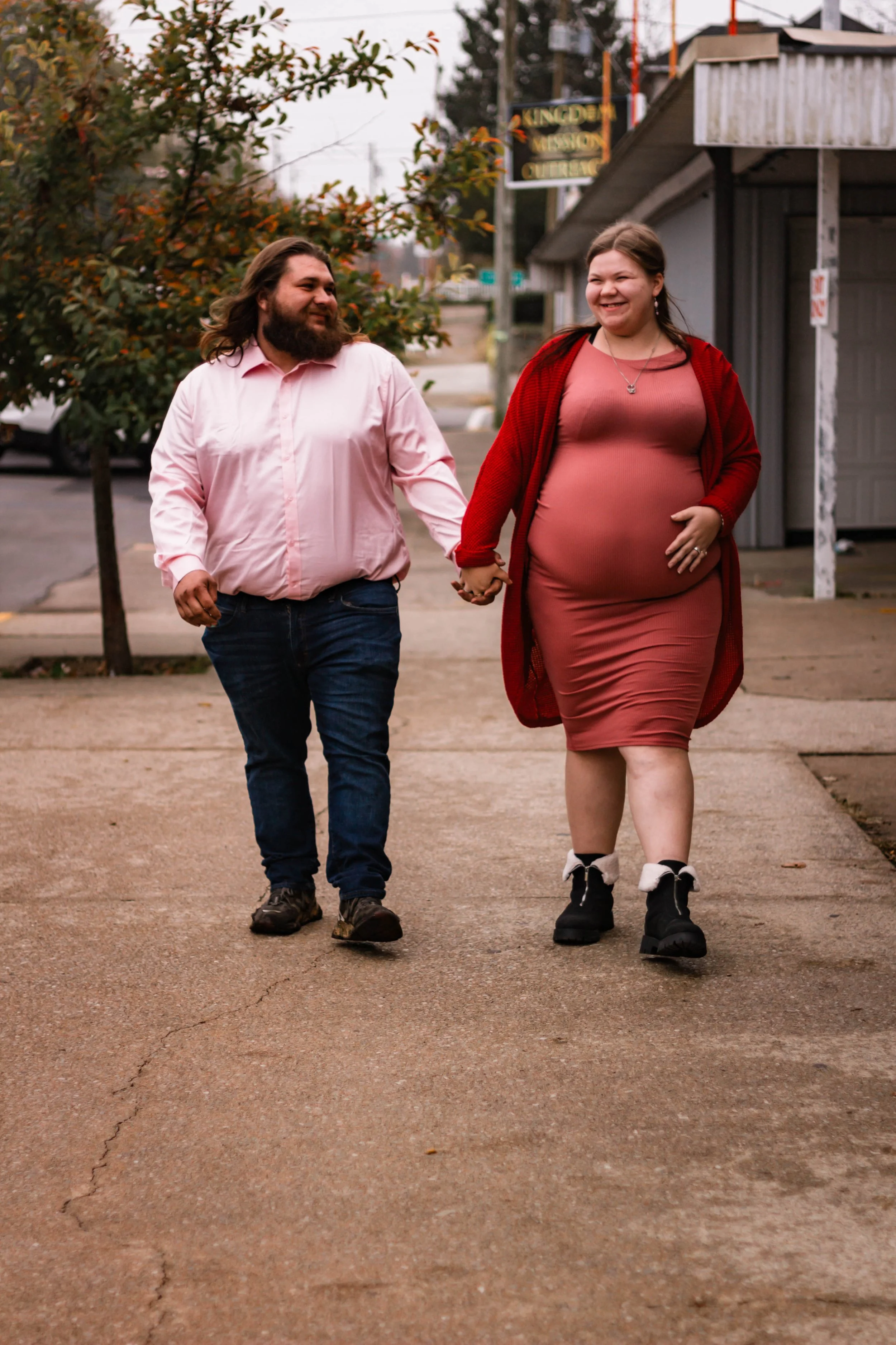 A man and a pregnant woman walking hand in hand on a sidewalk in an urban area, smiling and looking at each other.